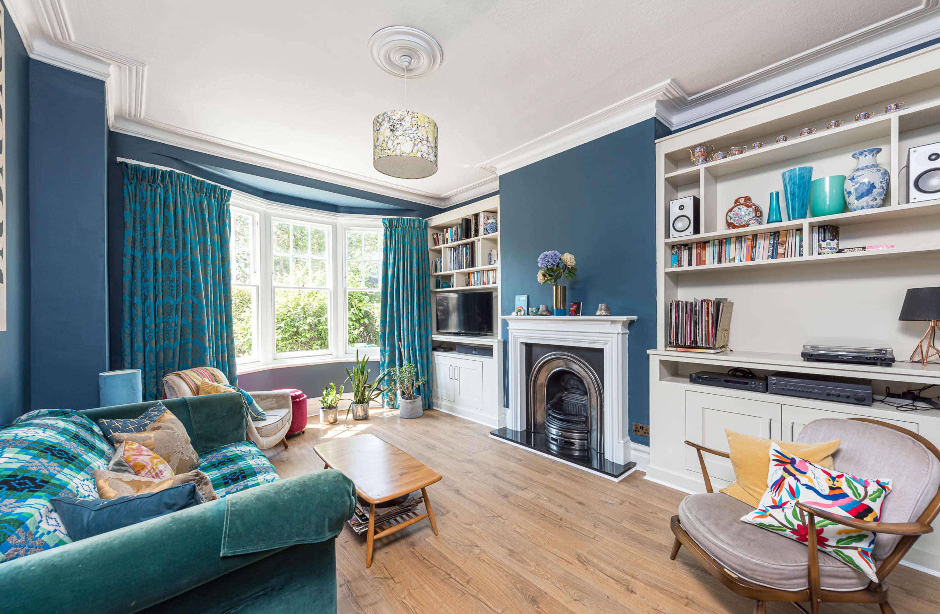 The image shows a well-lit living room with a bay window, shelves filled with books and decorative items, a cozy sofa, and a fireplace.
