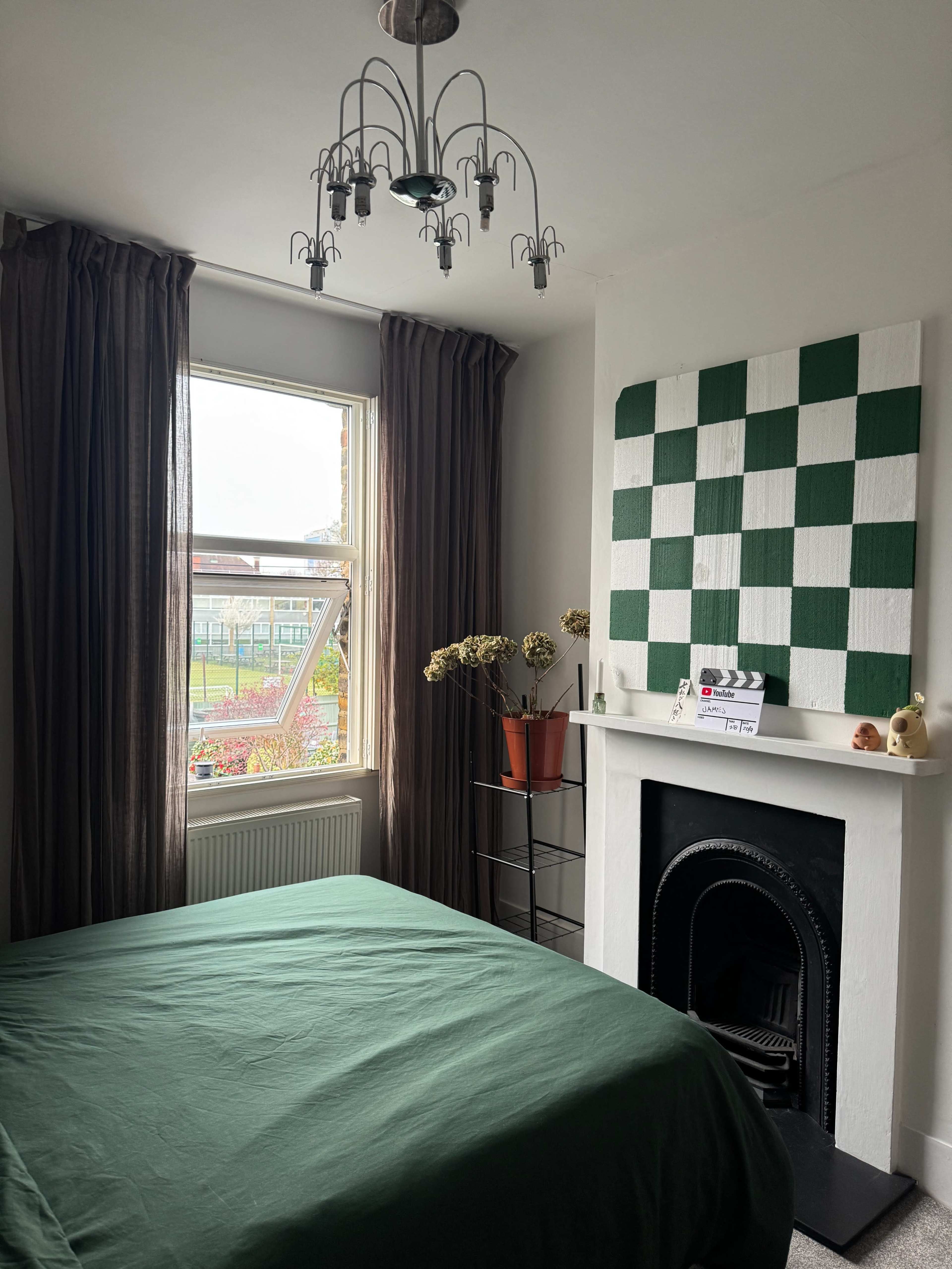 The image shows a bedroom featuring a green bedspread, a window with brown curtains, and a decorative checked pattern on the wall above a fireplace.