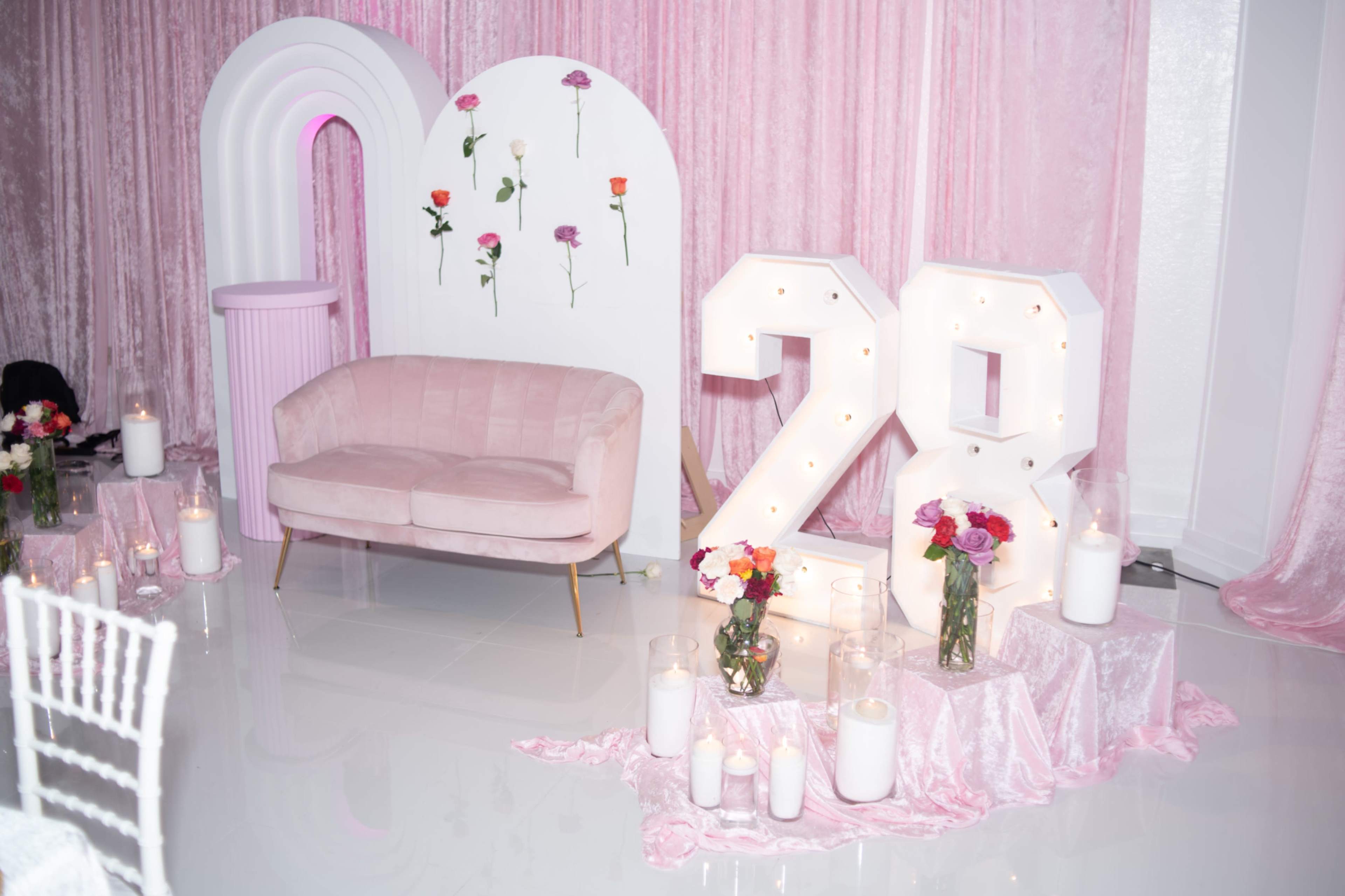 The image shows a decorated event space featuring a pink velvet sofa, large illuminated "28" numerals, and floral arrangements against a backdrop of pink draping.