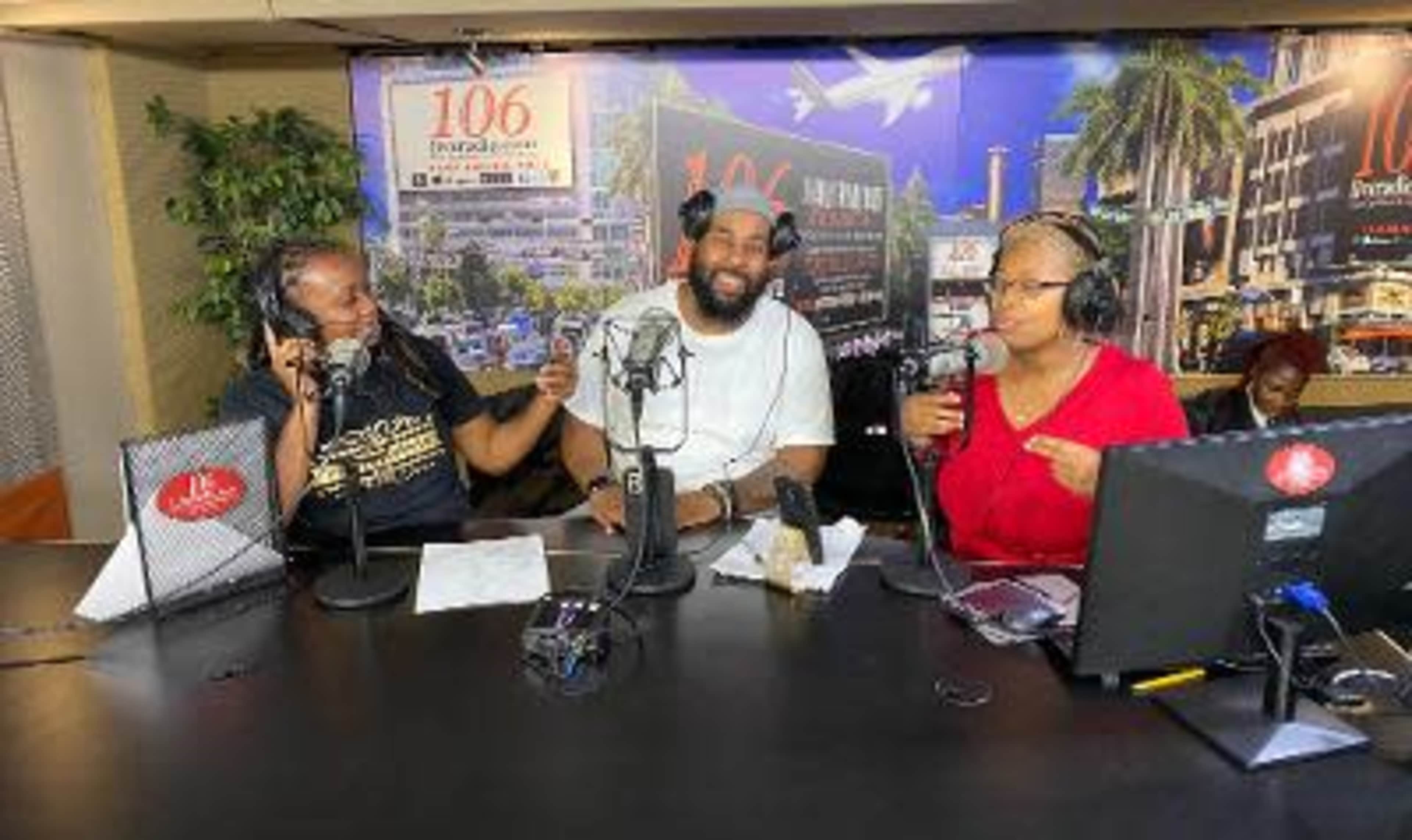 Three hosts are seated at a radio station desk, discussing topics while using microphones and wearing headphones, with a backdrop of a cityscape.