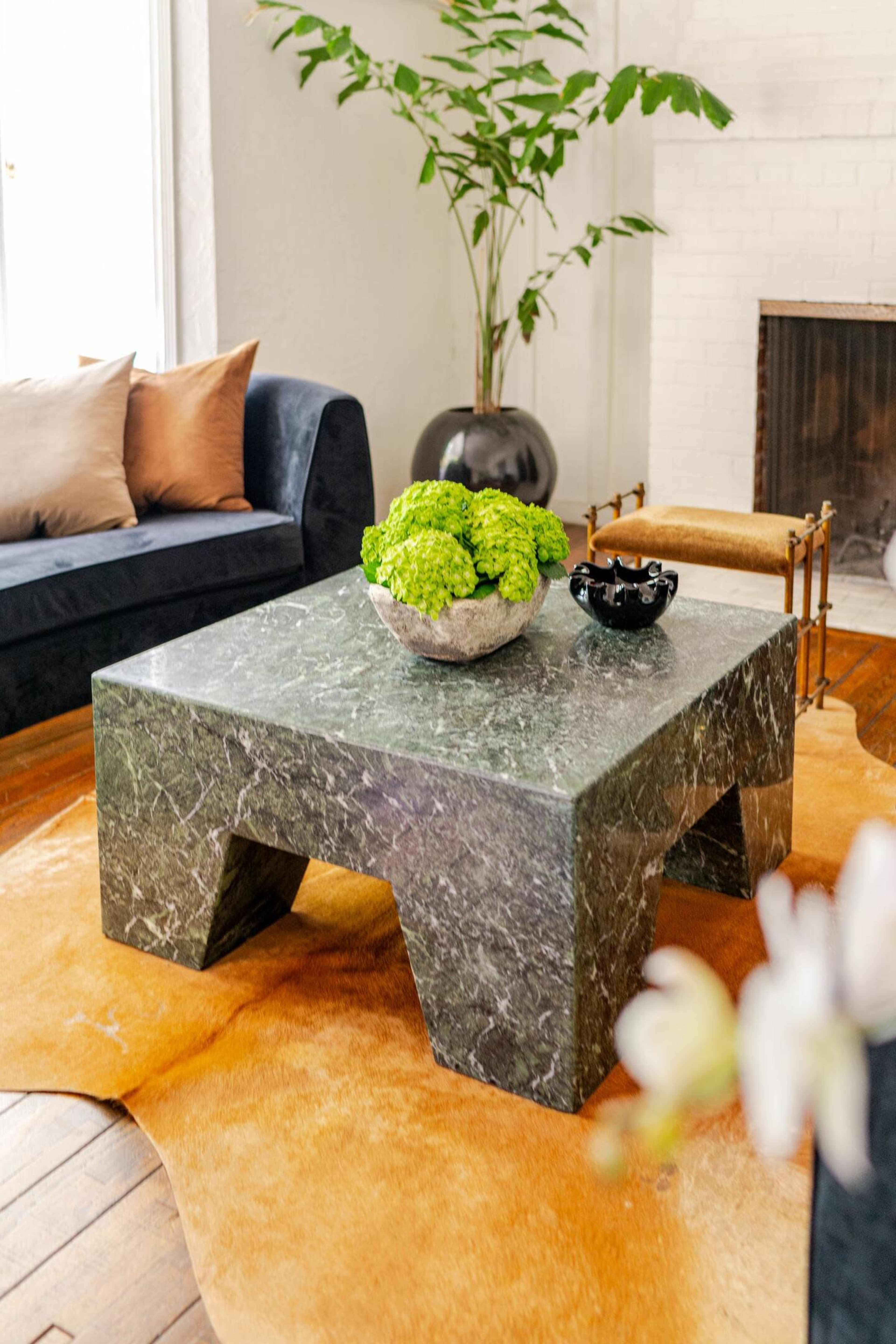 A green marble coffee table with a bowl of greenery sits on a tan rug in a living room equipped with a black sofa and a fireplace.
