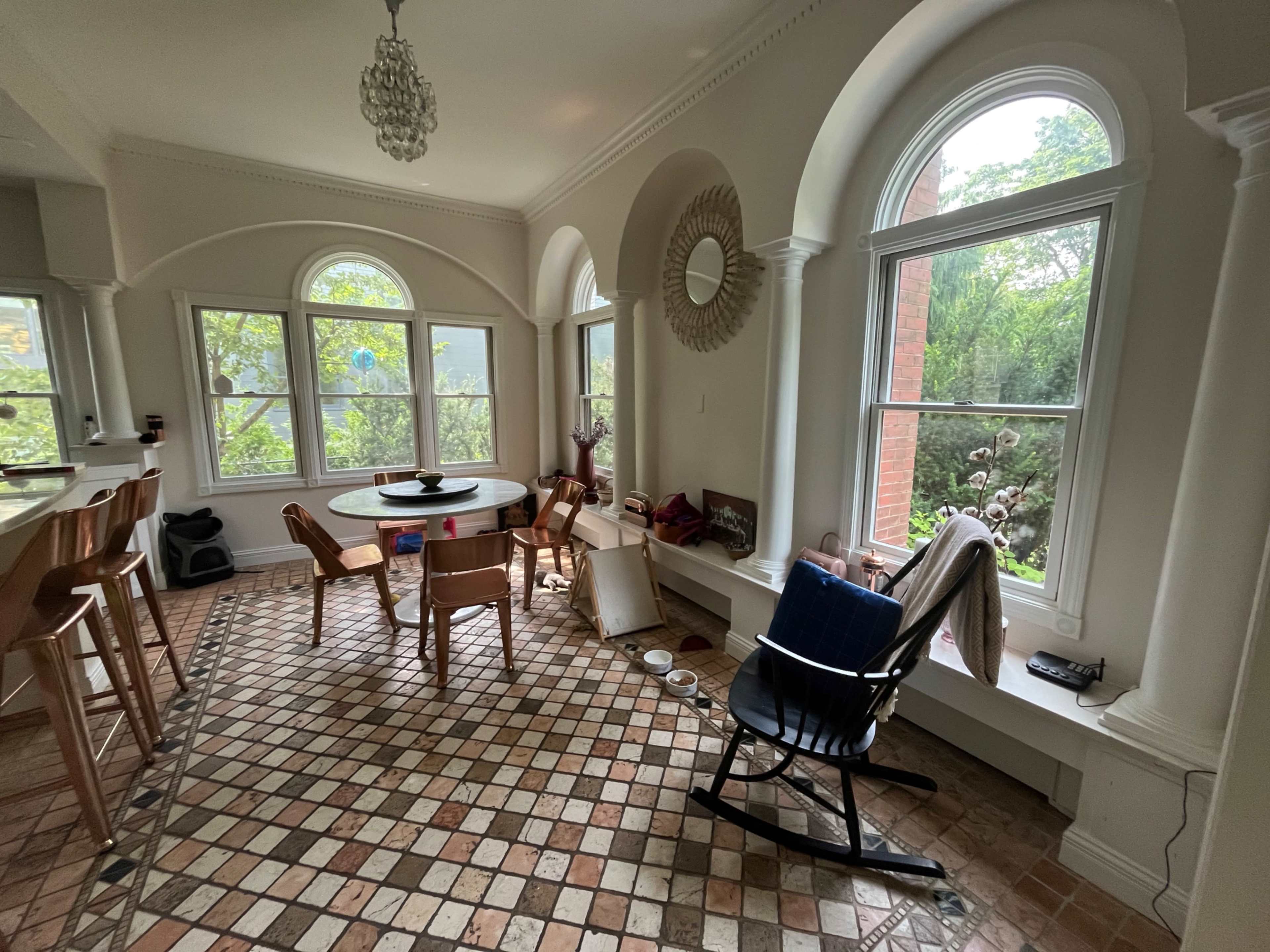 A bright dining area features a round table surrounded by wooden chairs, large windows providing a view of greenery, and a patterned tiled floor.