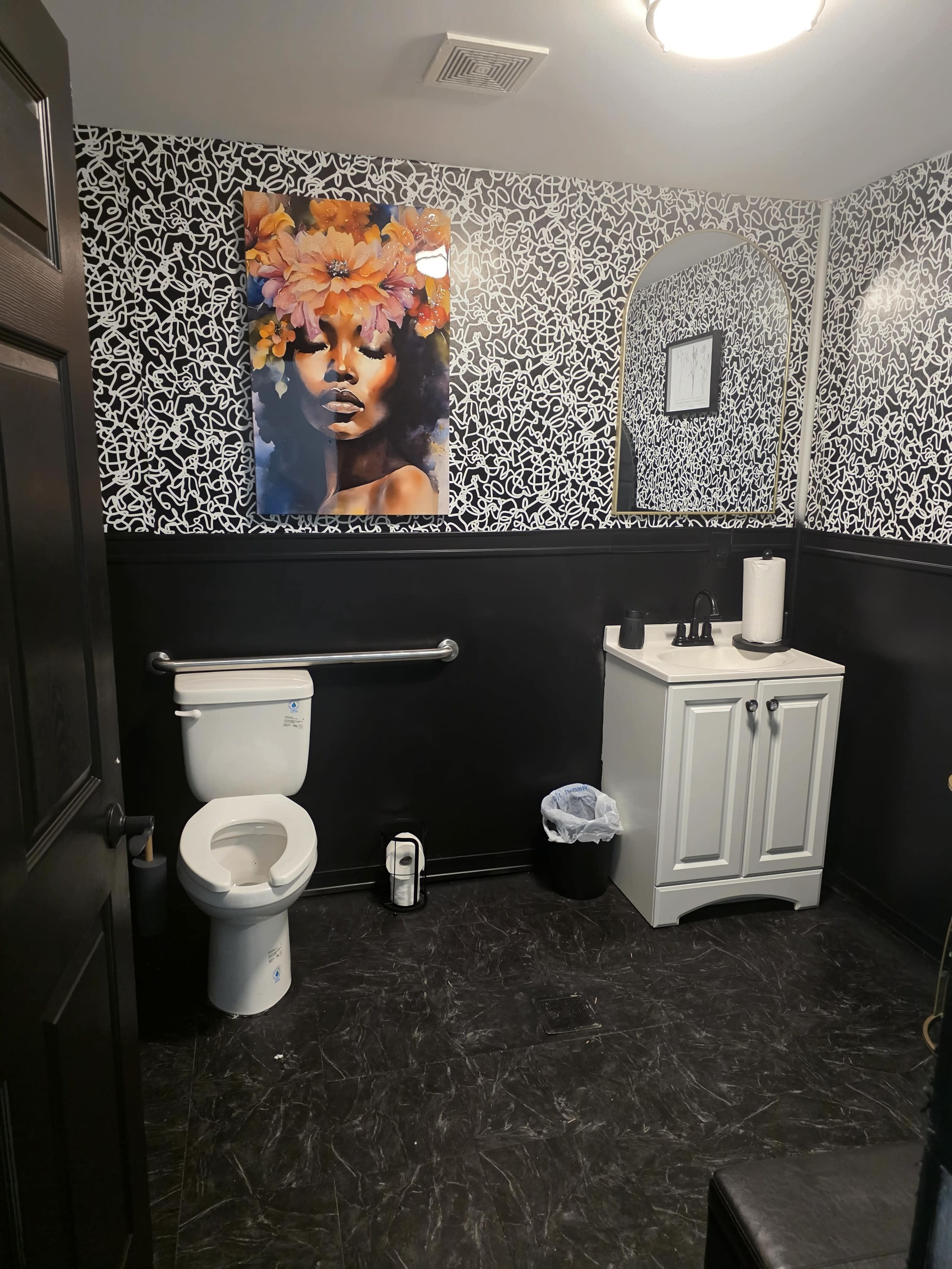 The bathroom features a black and white floral wall design, a mural of a woman with flowers in her hair, a toilet, a small vanity with a sink, and a wastebasket.