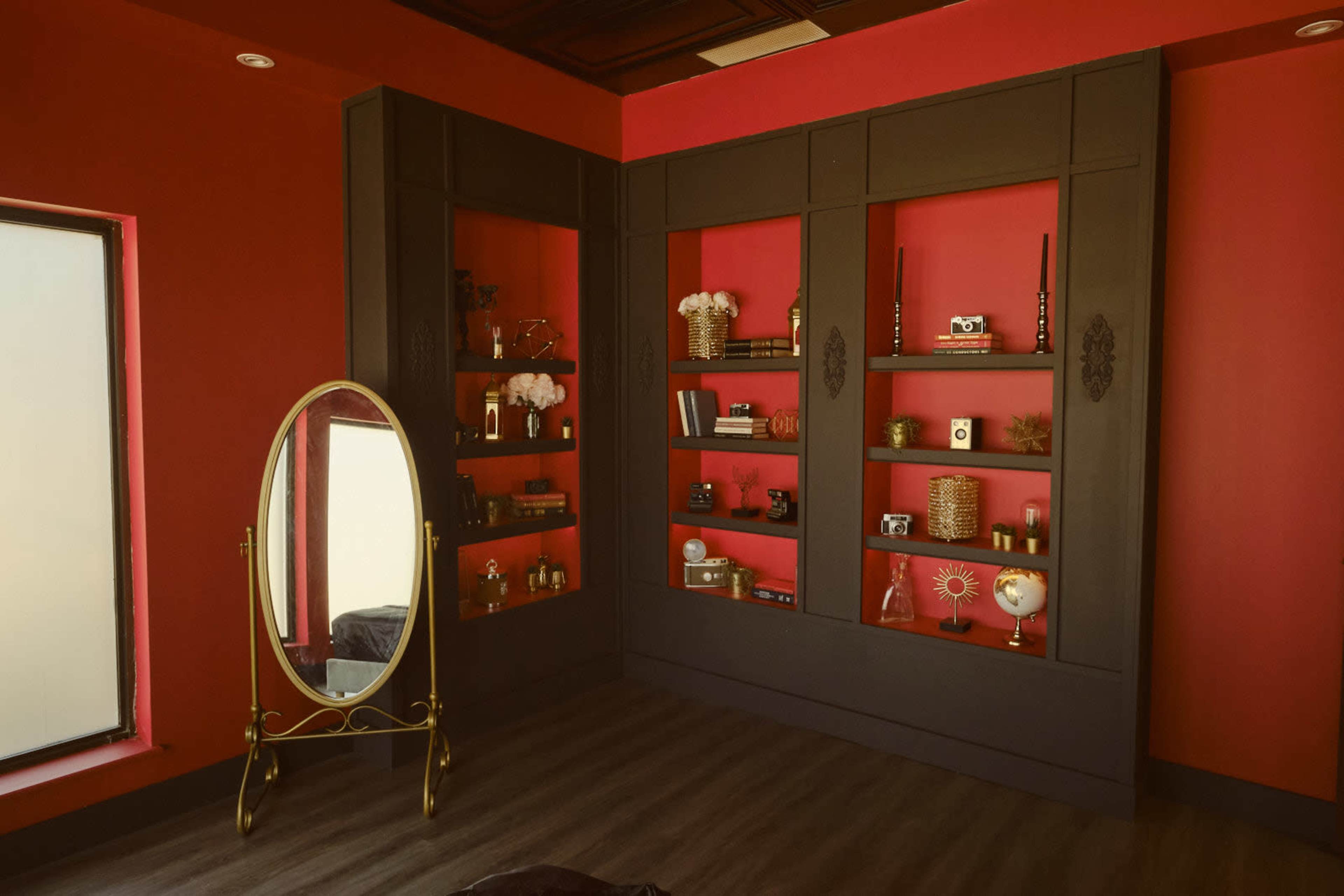 The image shows a room with red walls featuring a dark cabinetry unit filled with decorative items and a gold-framed mirror standing on the floor.