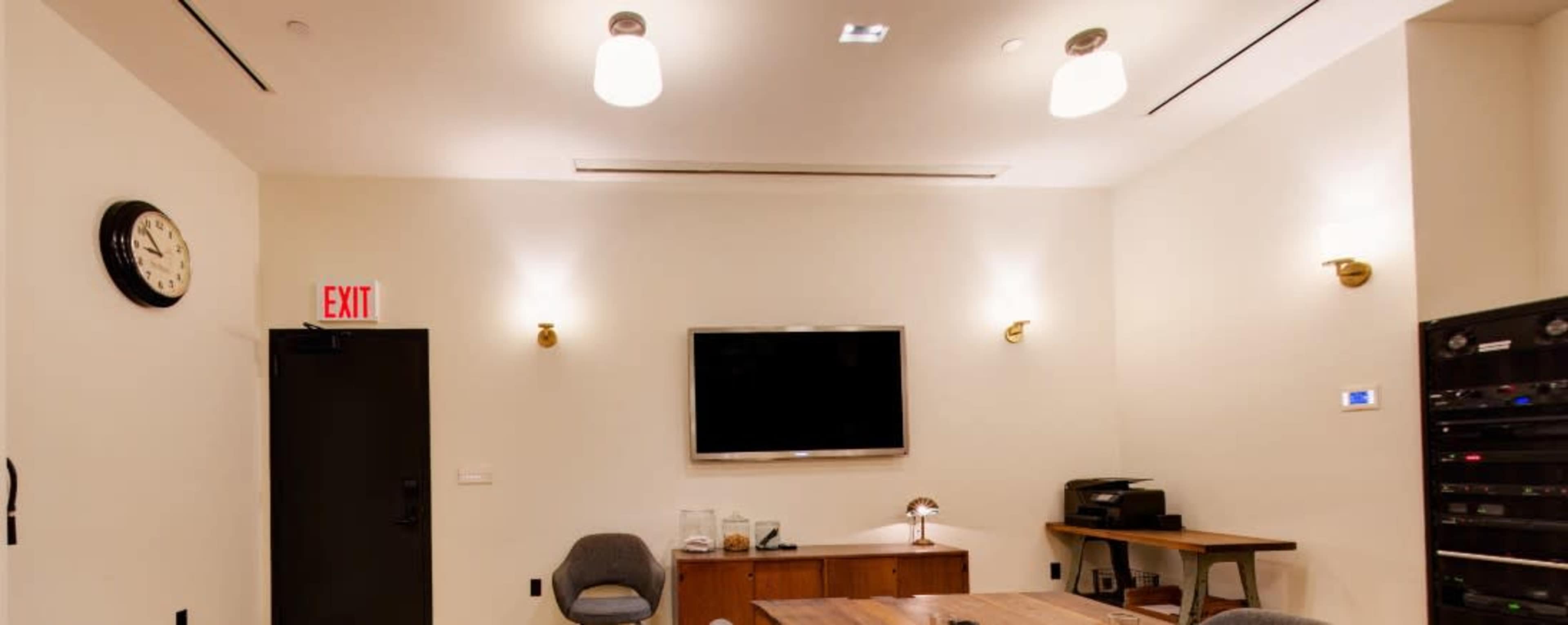 A conference room features a wall-mounted television, a wooden table with chairs, and a clock on the wall.