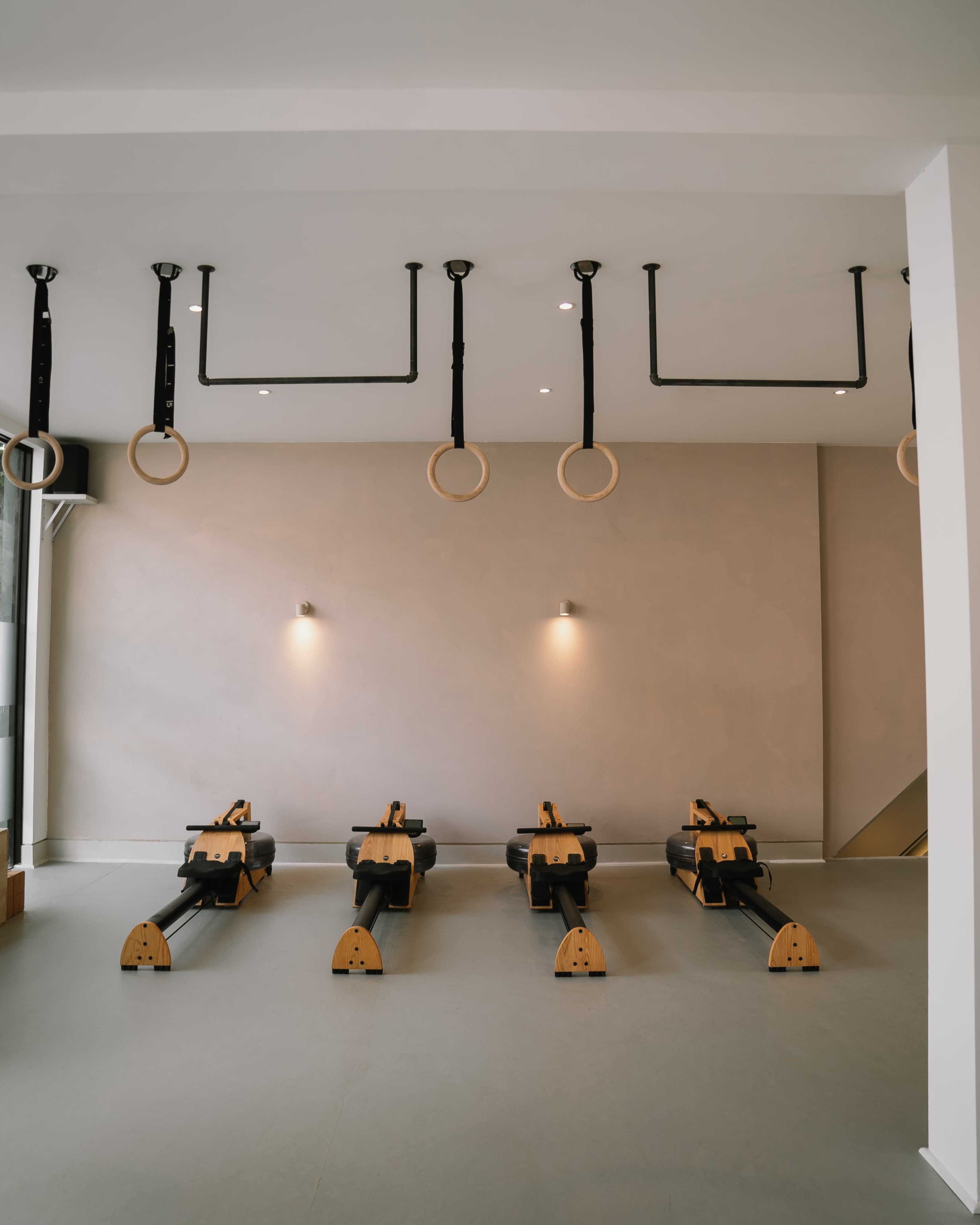 A minimalist gym interior featuring four rowing machines in a row, with gymnastic rings hanging from the ceiling.