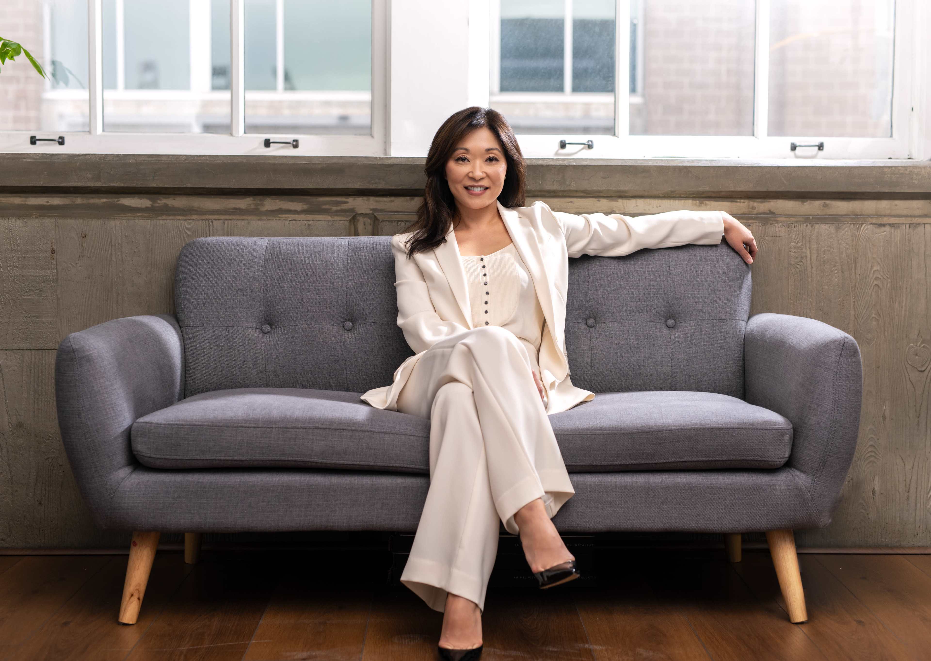 A woman in a white suit sits confidently on a gray sofa in a well-lit room with large windows.