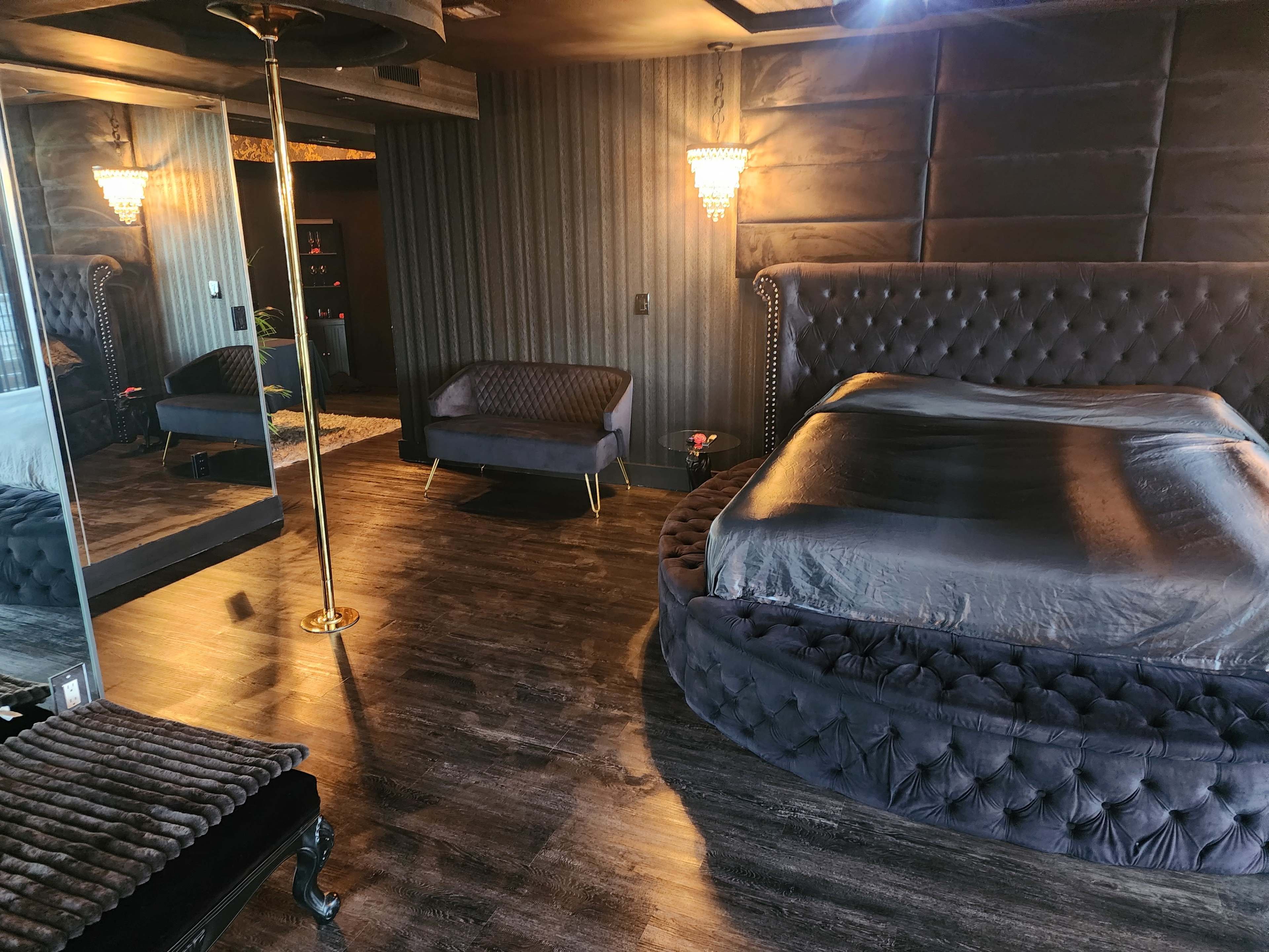 The room features a large, round bed with a velvet tufted headboard, a mirrored wall, and a pole beside a small lounge chair.