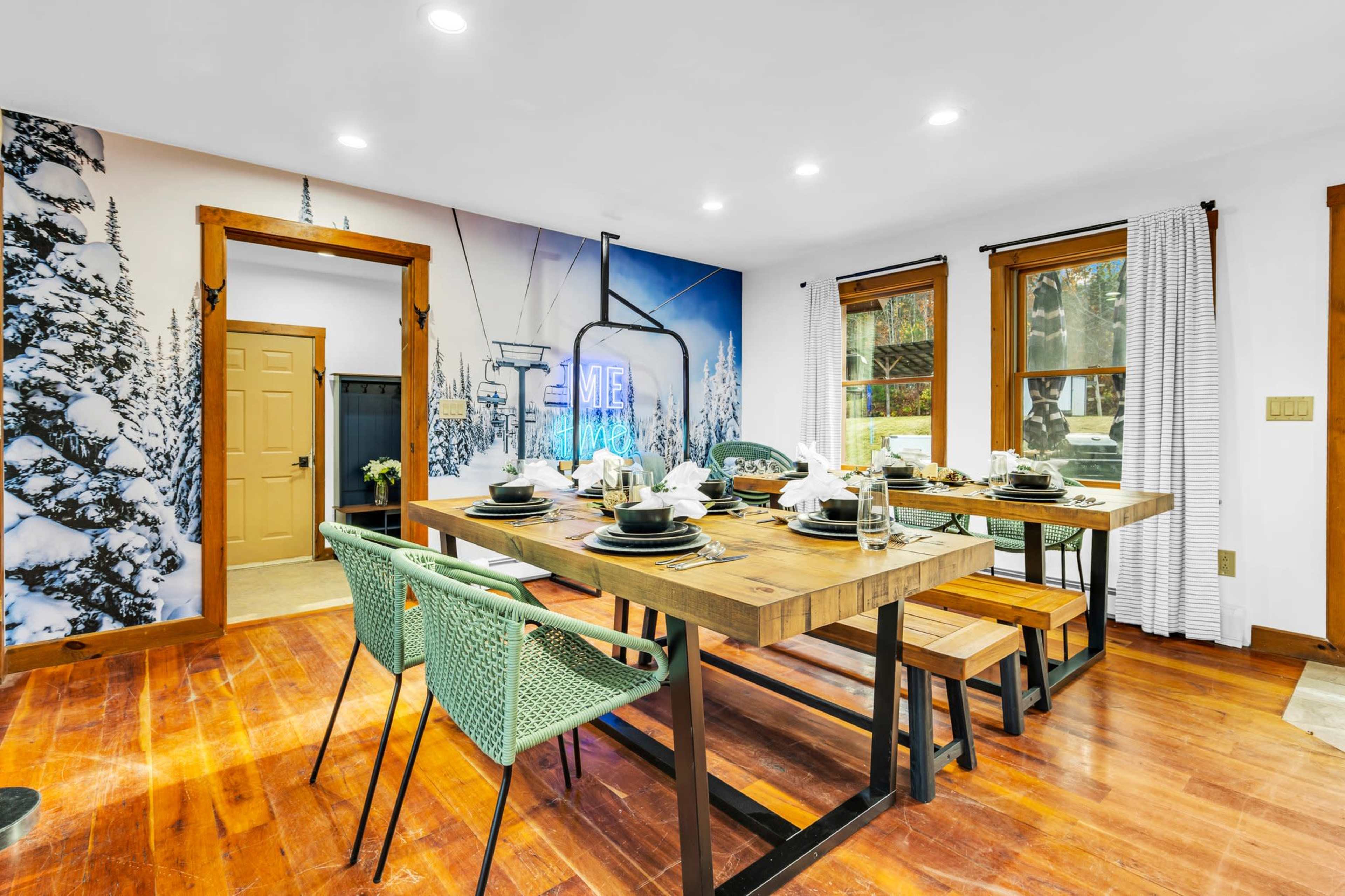 A dining area features a wooden table set for a meal, surrounded by green chairs, with a backdrop of a mural depicting snow-covered trees and a ski lift.