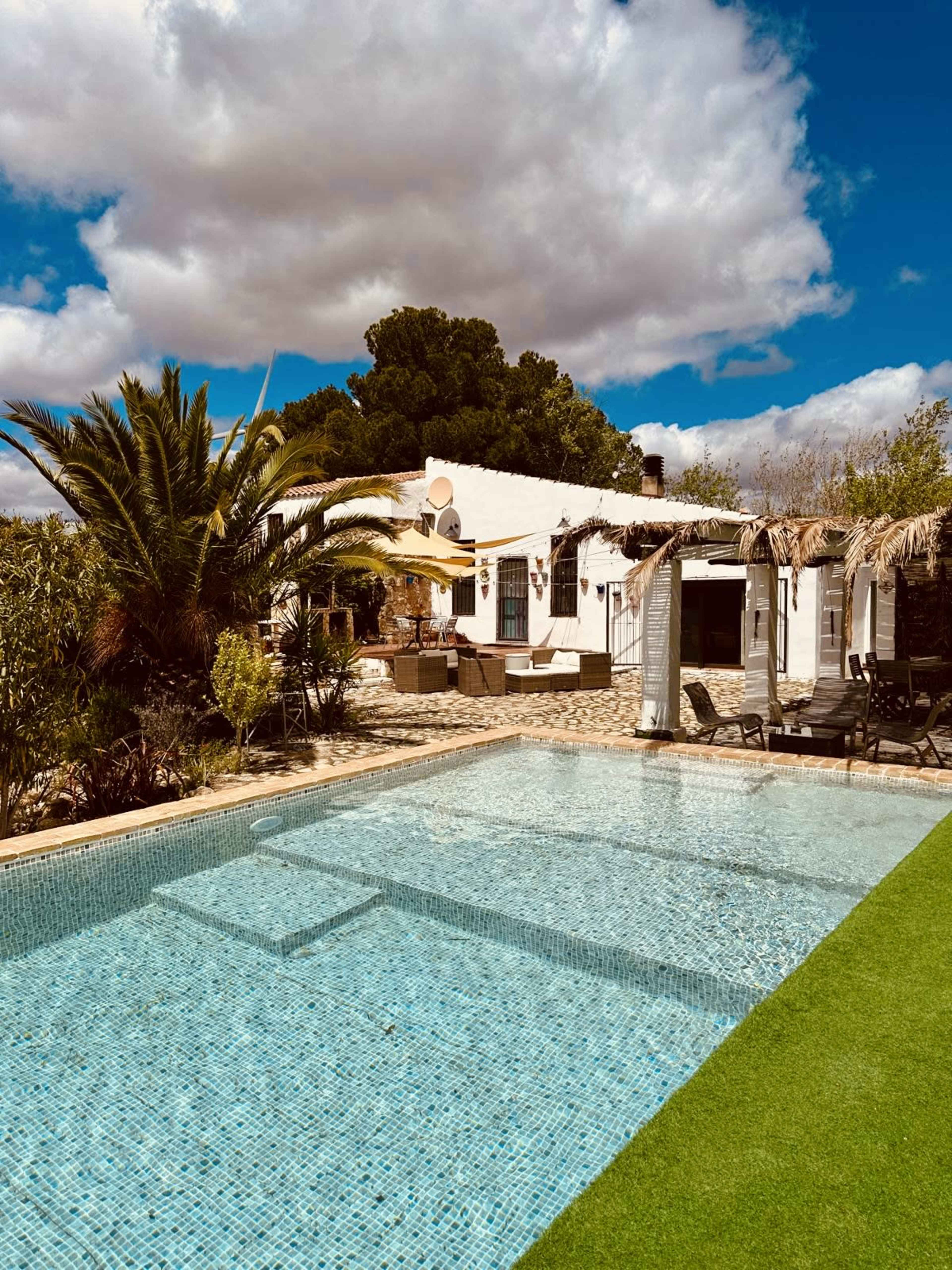 A clear swimming pool reflects the sky in front of a white house surrounded by greenery and lounging areas.
