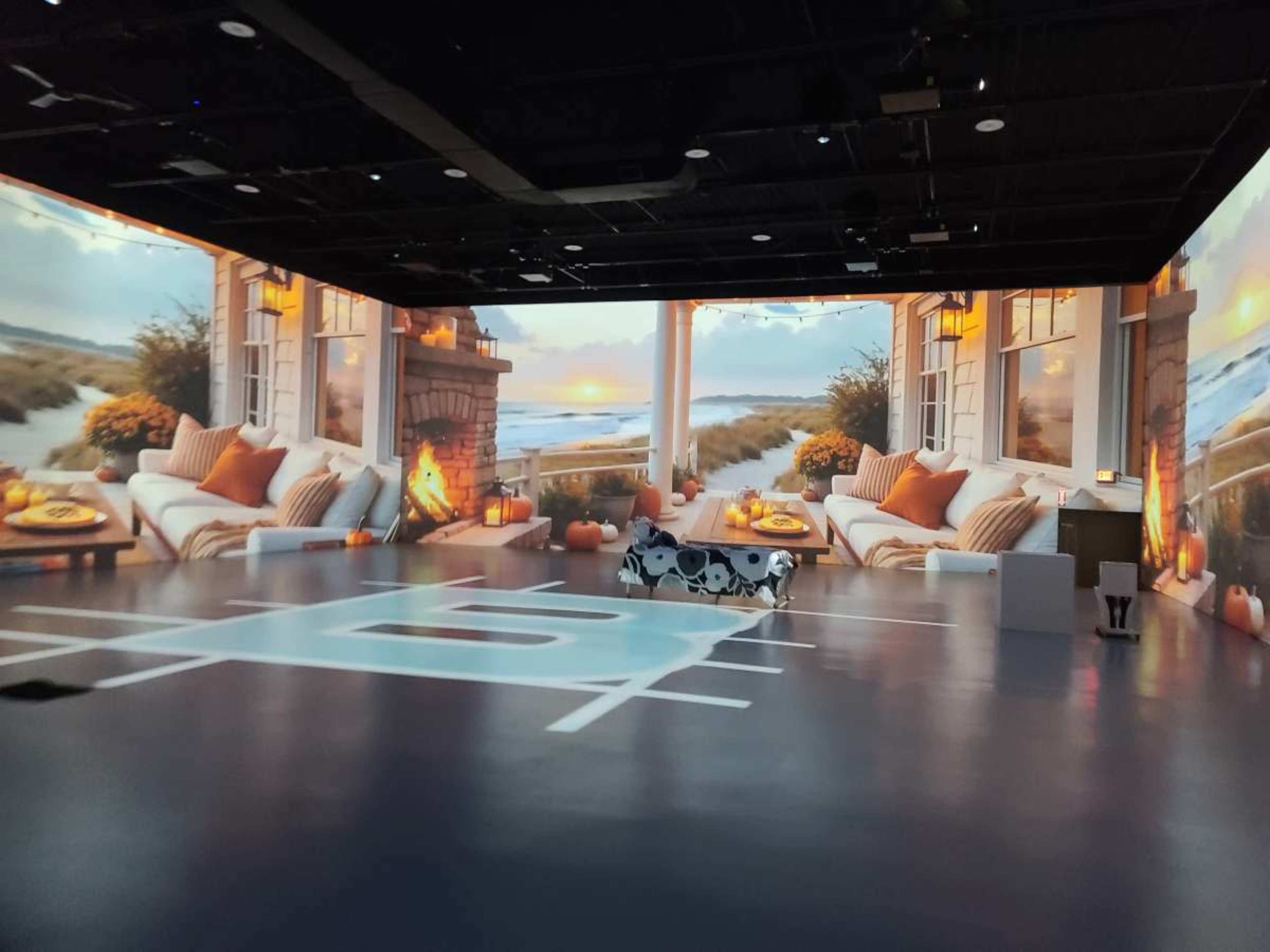 The image shows a large room with immersive wall projections depicting a cozy beachside living area at sunset, complete with comfortable seating and decorative elements.