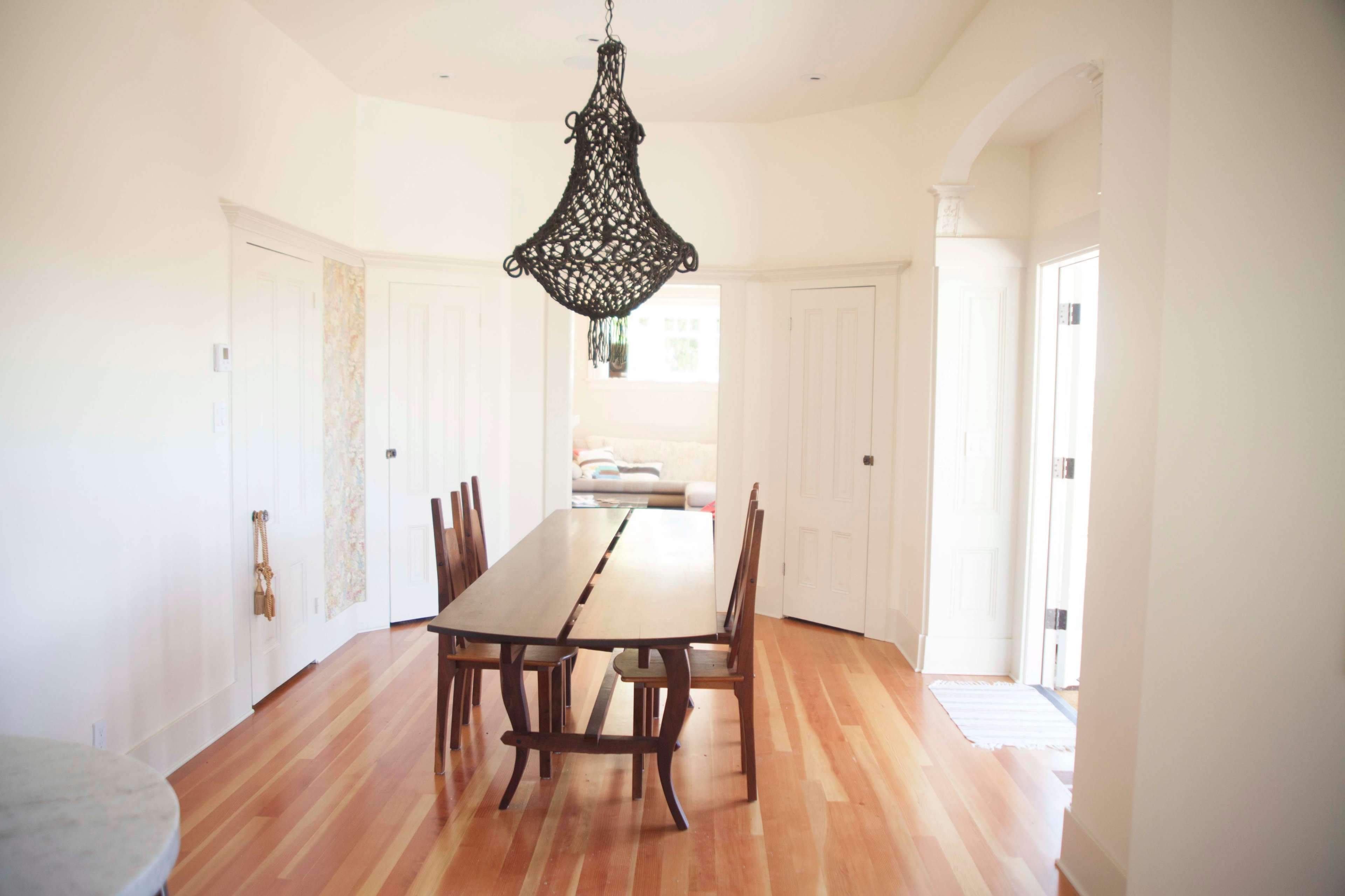A wooden dining table with four chairs is positioned in a bright, airy room with white walls and wooden flooring, flanked by two doorways.