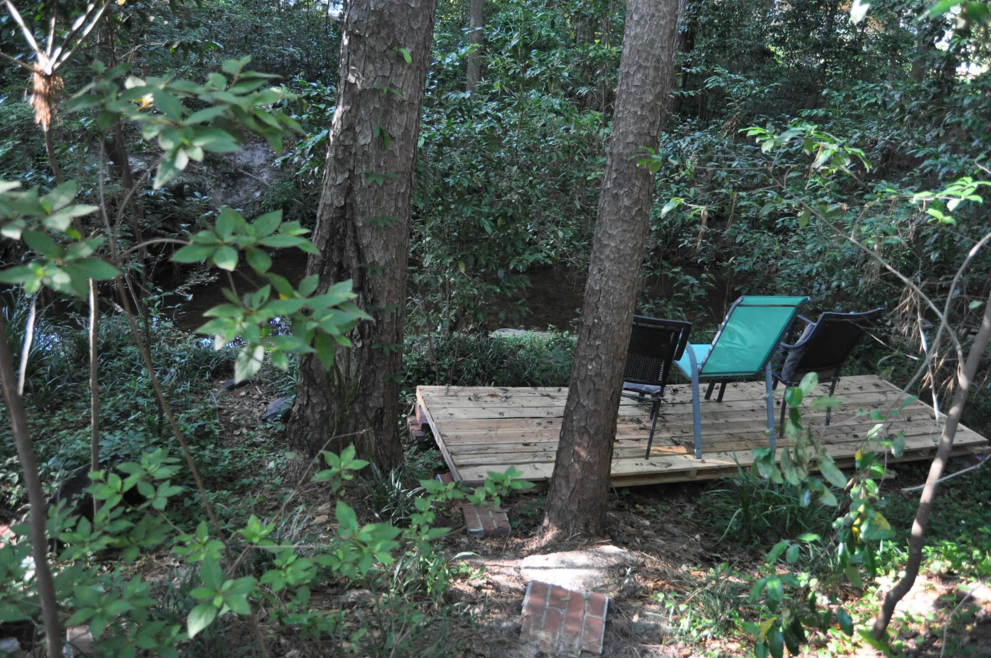 A wooden platform by a creek is adorned with two chairs nestled among trees and shrubs.