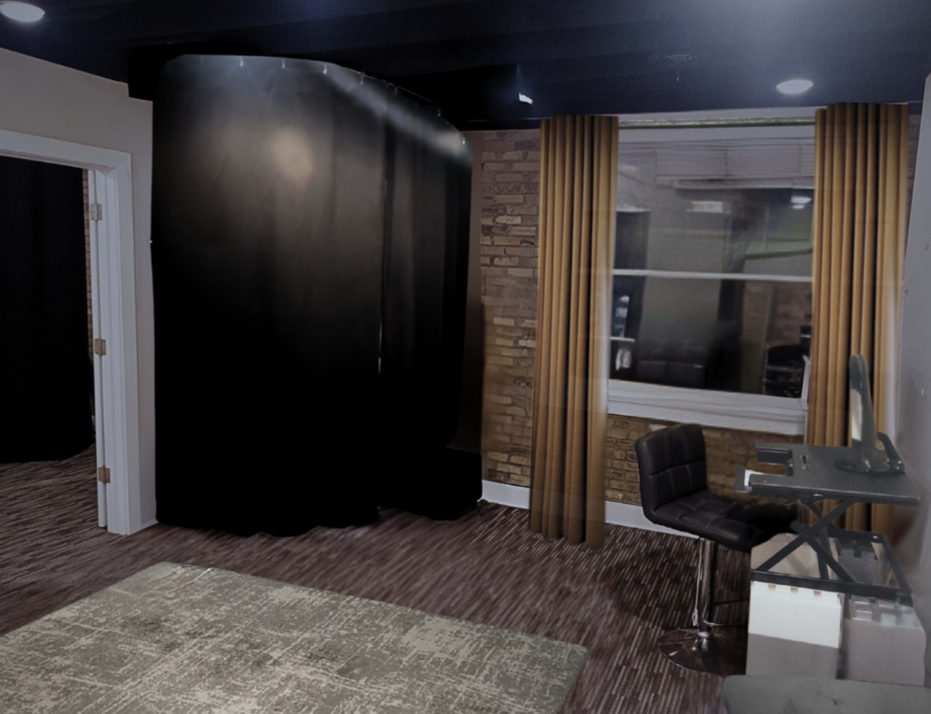 The image shows a room with a black curtain backdrop, brick walls, a window with curtains, and a small desk with a chair.