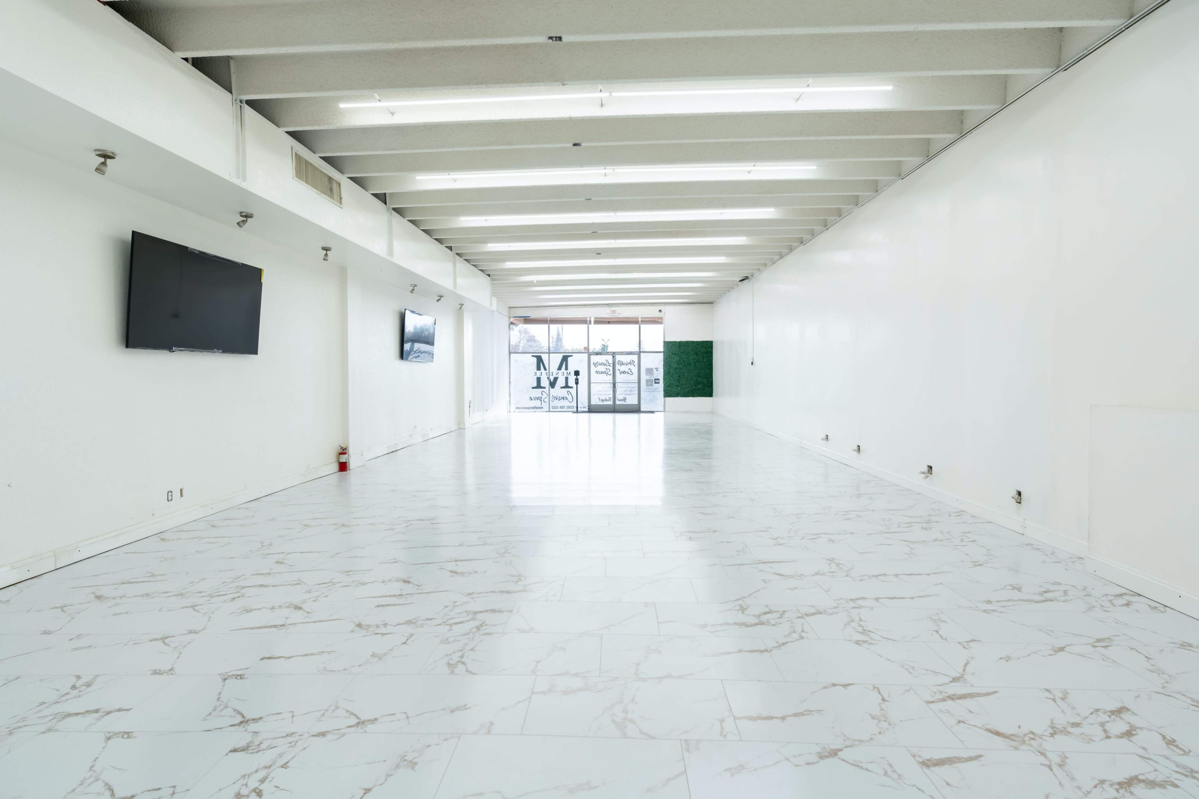 A vacant interior space with white walls, marble-patterned flooring, and two wall-mounted televisions.