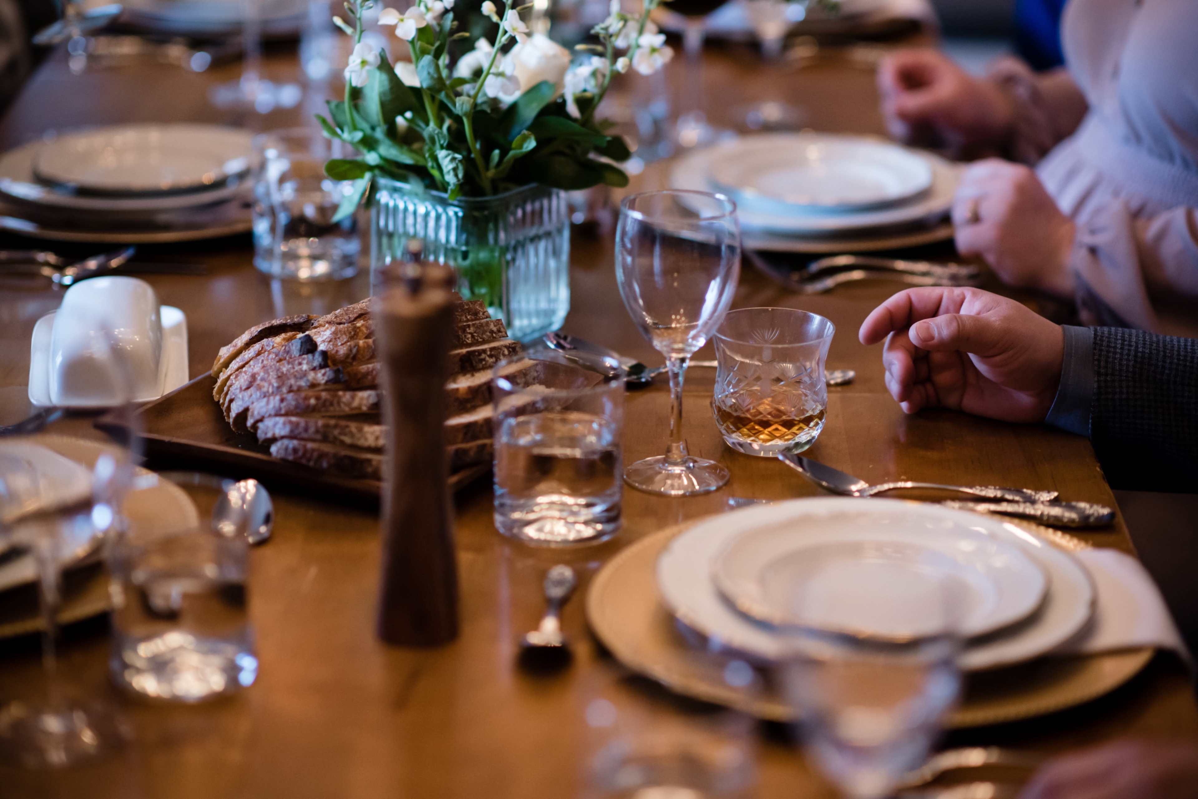 A dining table is set with plates, glasses, and a centerpiece of flowers, accompanied by a basket of bread and a bottle of whiskey.