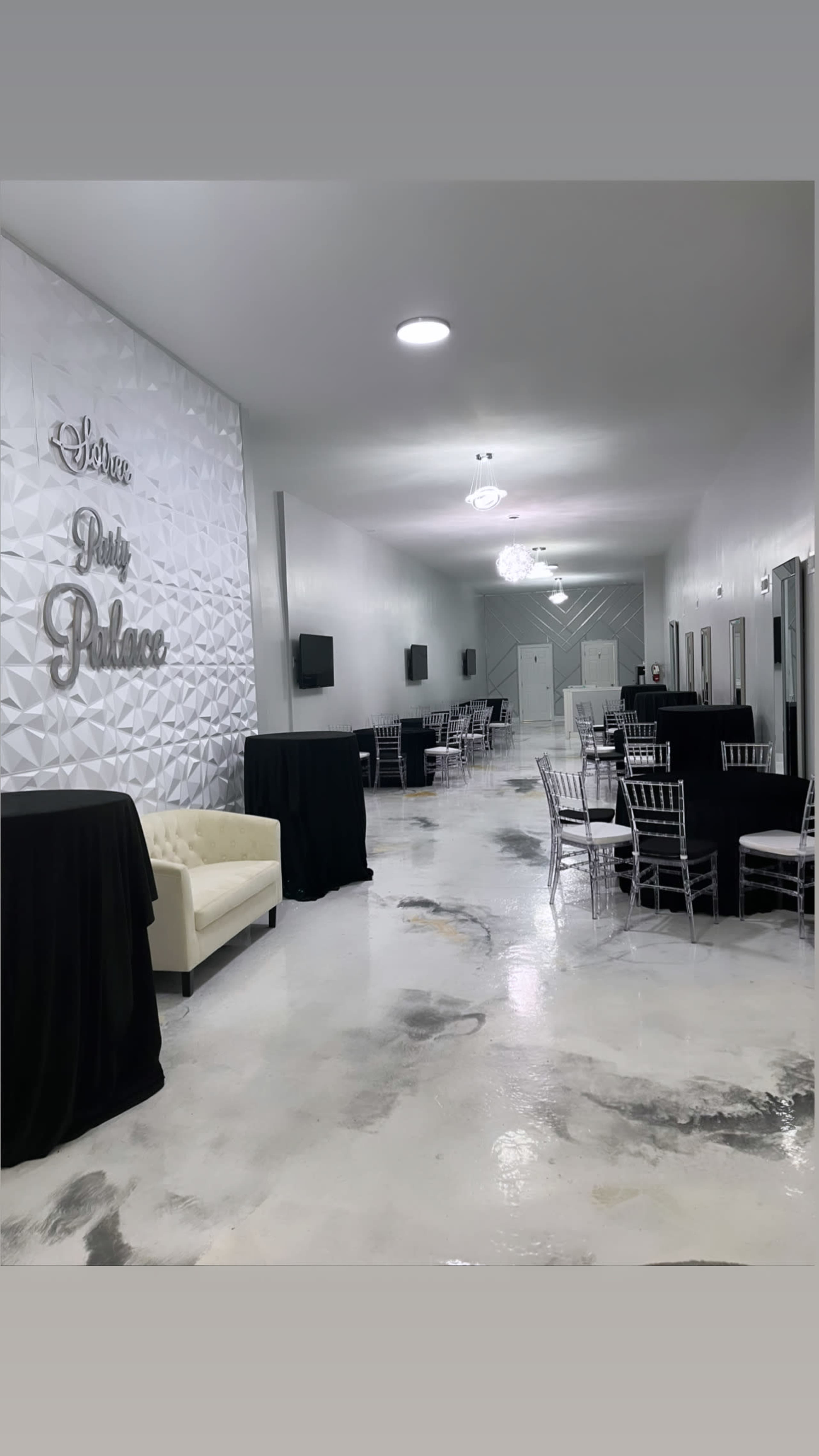 A spacious, well-lit event hall features a white wall with decorative lettering, black tablecloths on tables, and silver chairs arranged throughout the room.