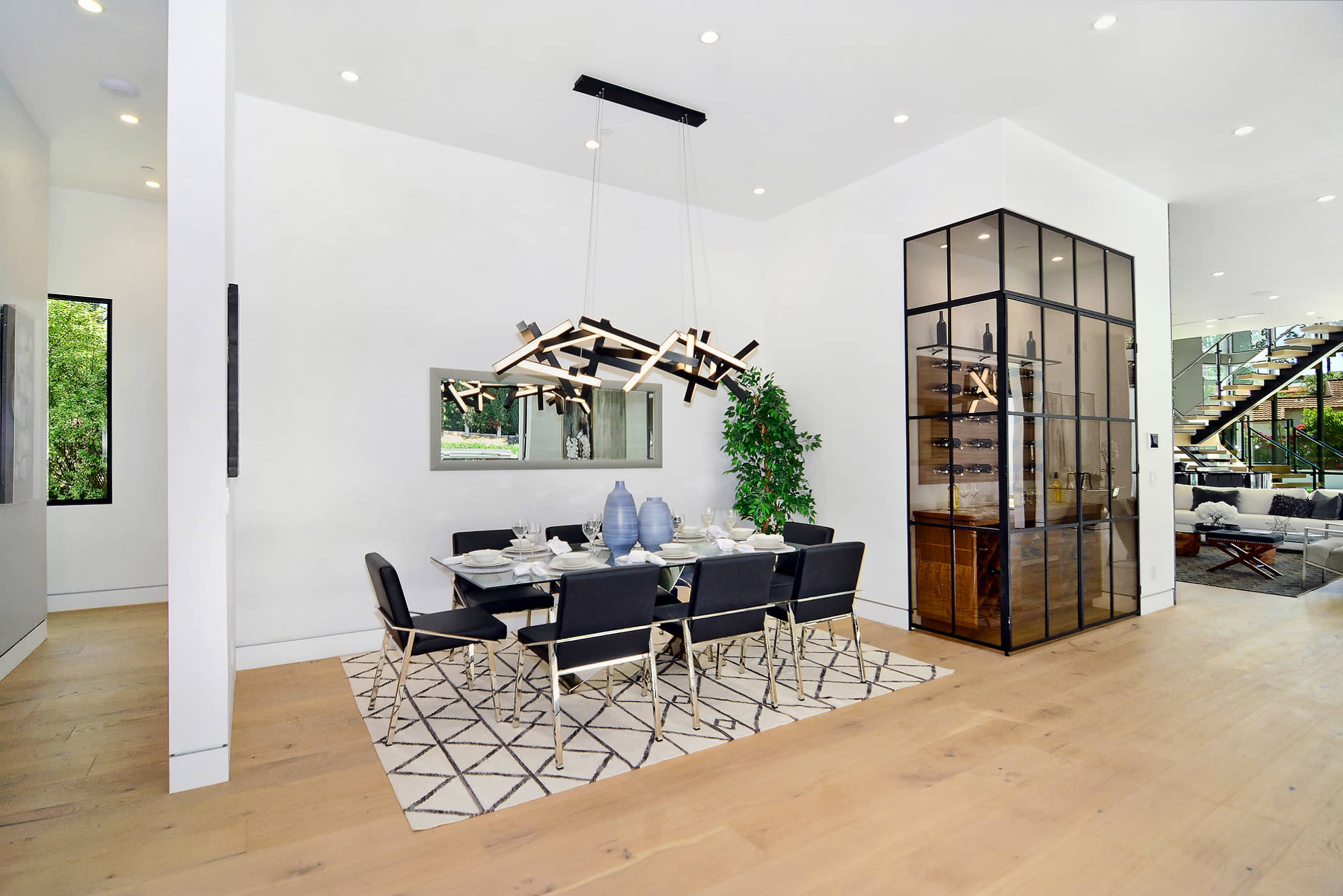 A modern dining area features a rectangular table with black chairs, a stylish light fixture above, and a glass cabinet in the background.