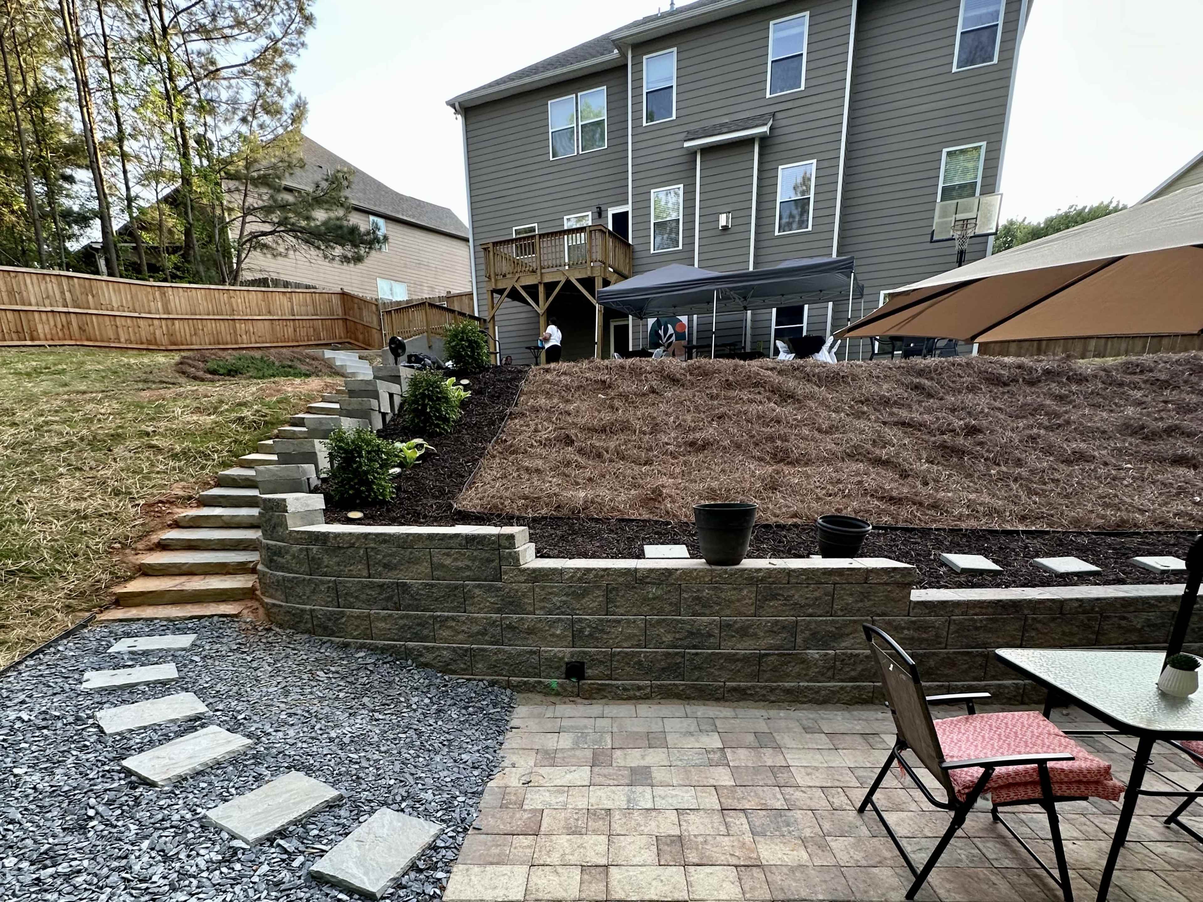 The image shows a landscaped backyard with a tiered area featuring mulch, decorative plants, a stone pathway, and a patio with outdoor furniture.