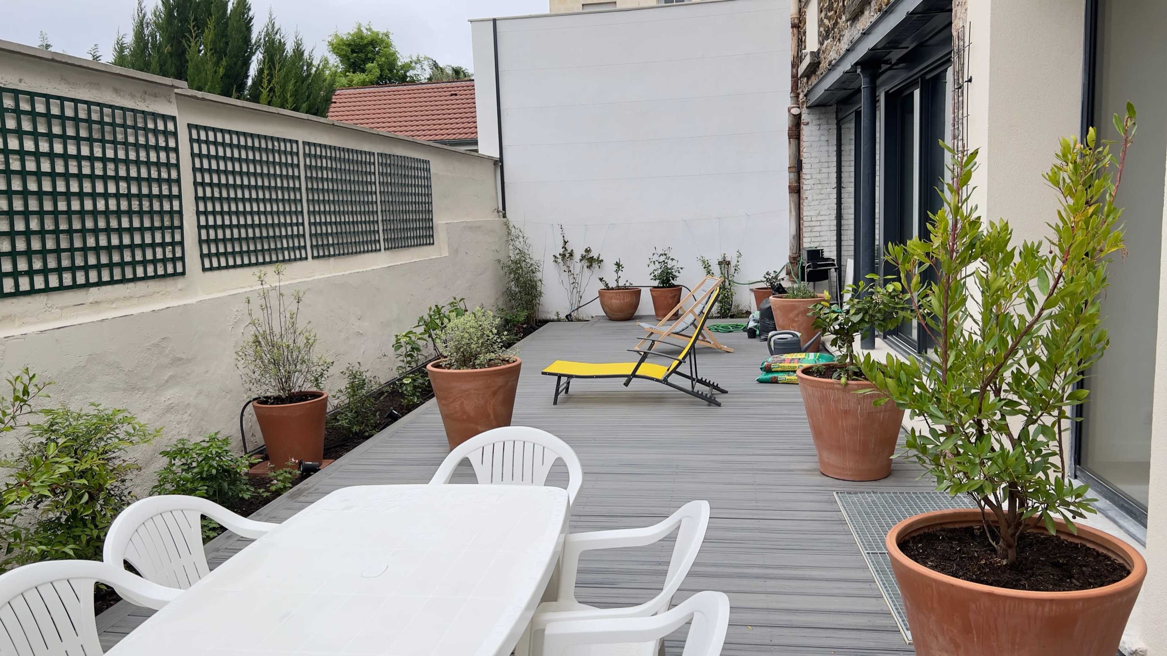 A wooden deck area with a table and white chairs, two loungers, and potted plants along the sides.