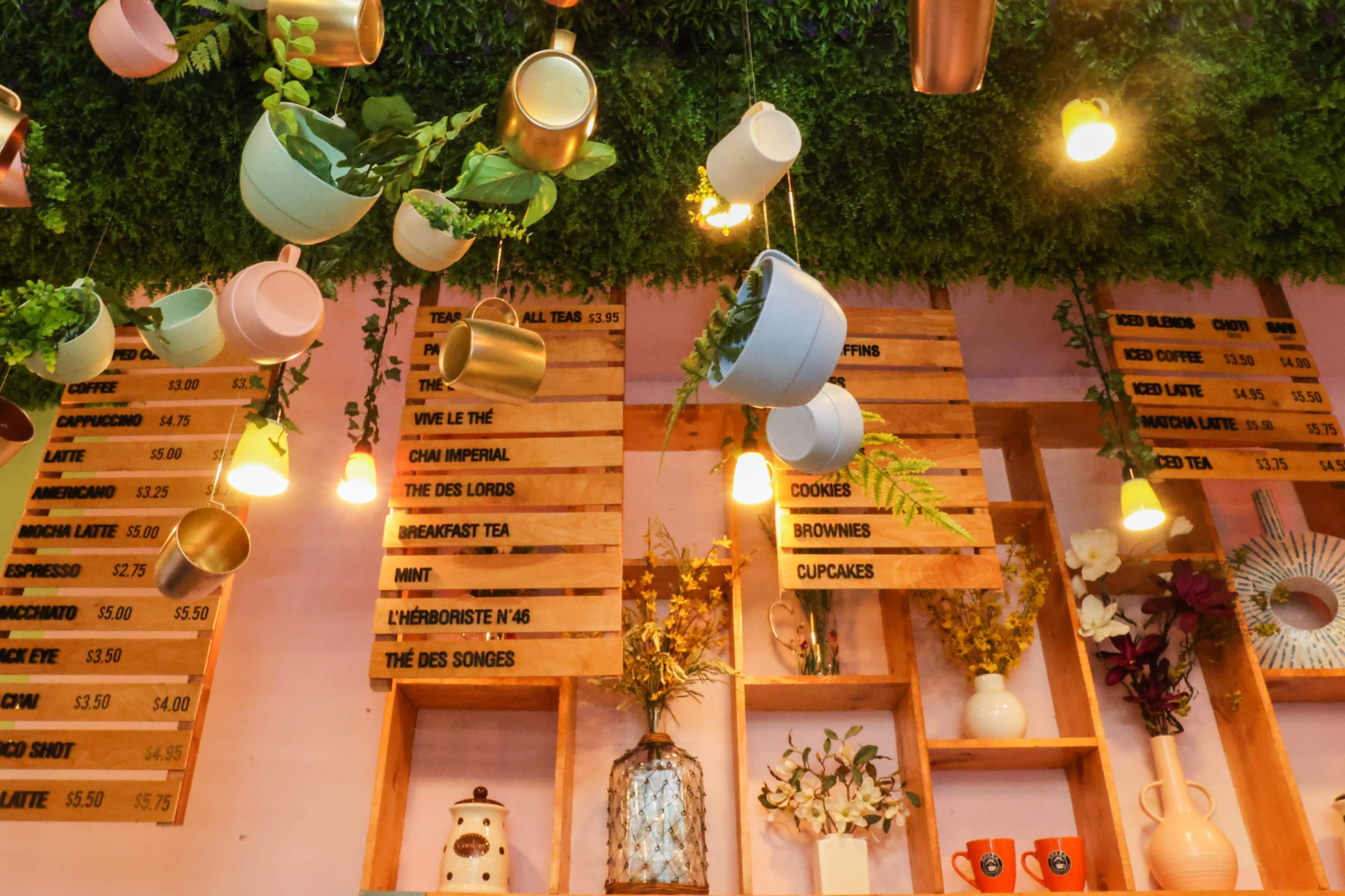 A café interior with hanging planters, illuminated light fixtures, and a wooden menu board displaying various beverages and snacks.
