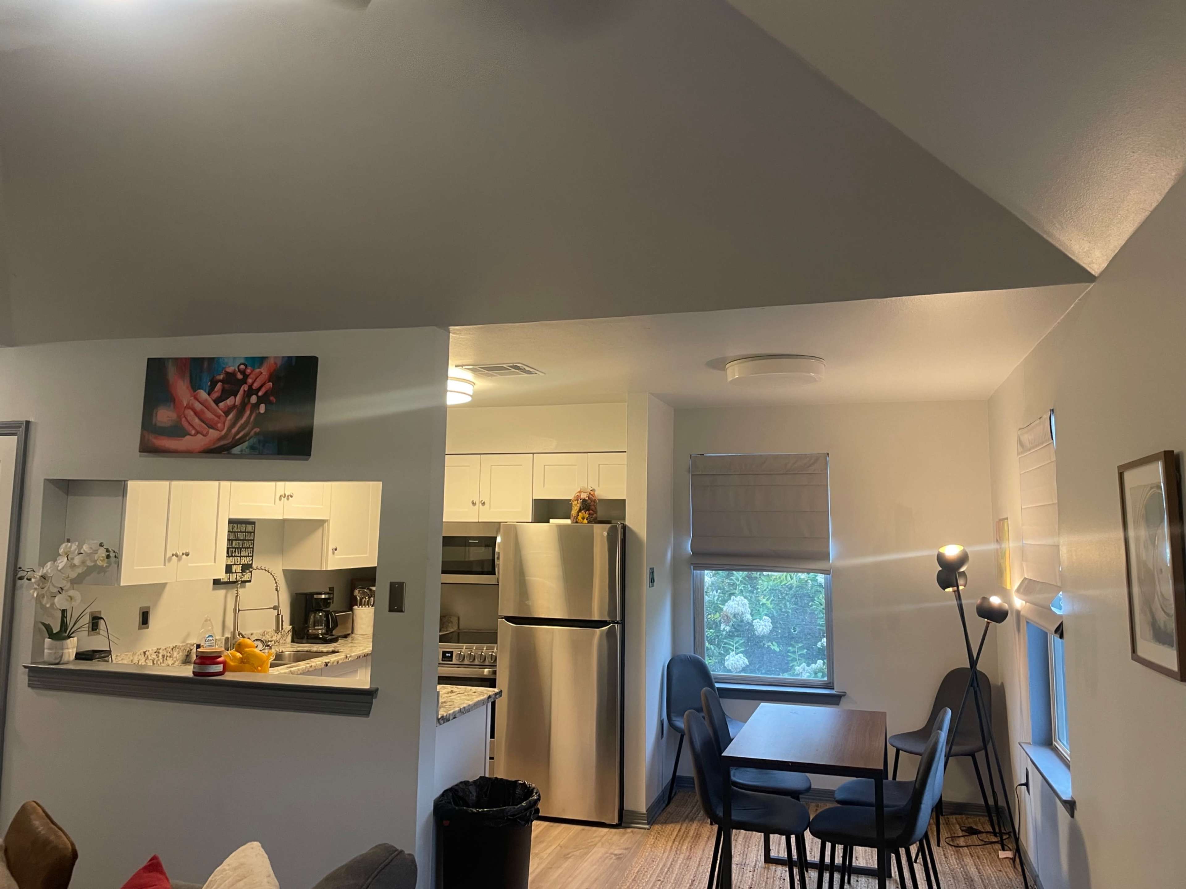The image shows a modern kitchen and dining area with a table and chairs, a stainless steel refrigerator, and a decorative artwork on the wall.