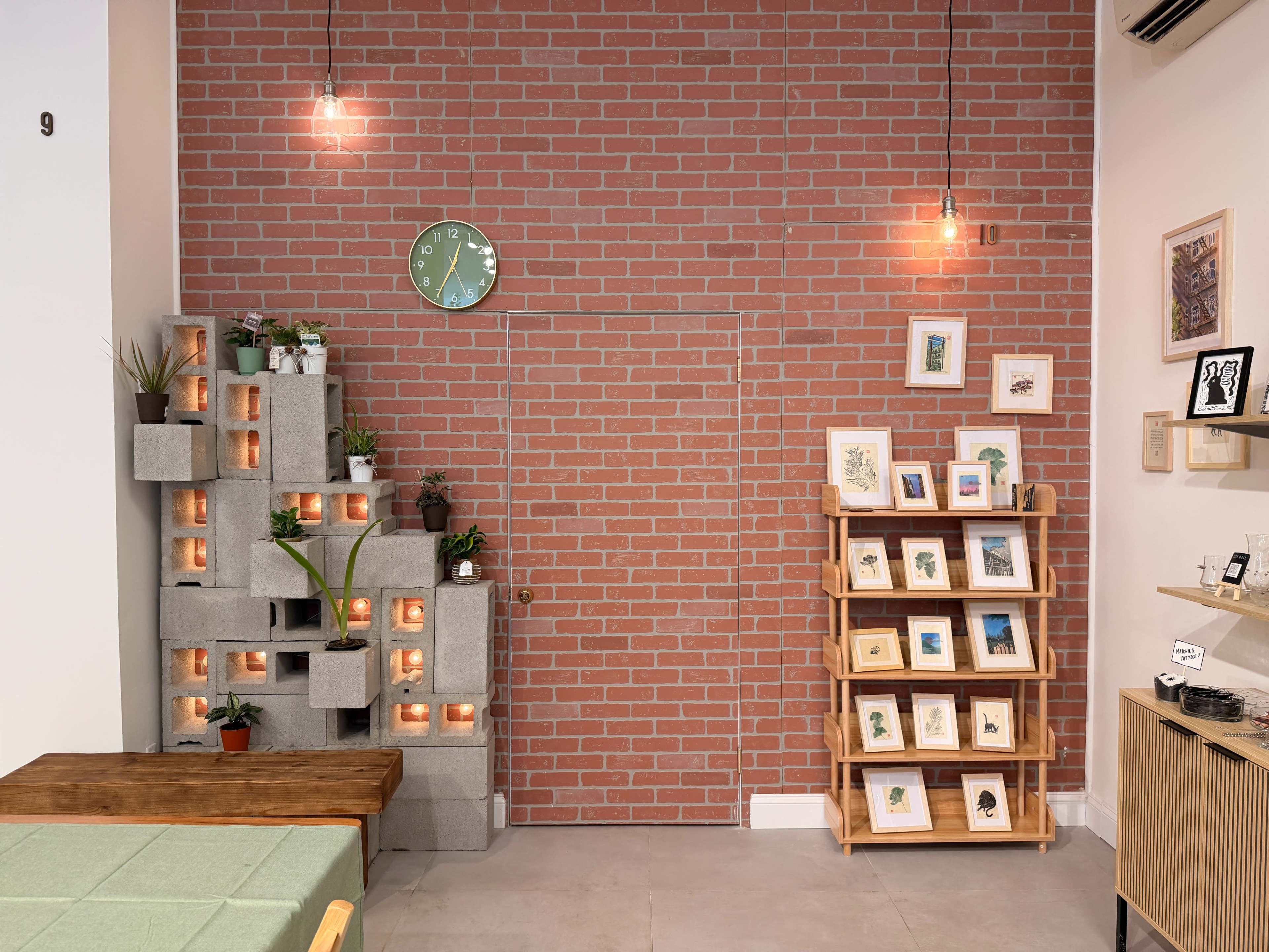 The image shows a modern cafe interior featuring a brick wall, a decorative concrete block planter, a wall clock, framed art on display, and a wooden table.