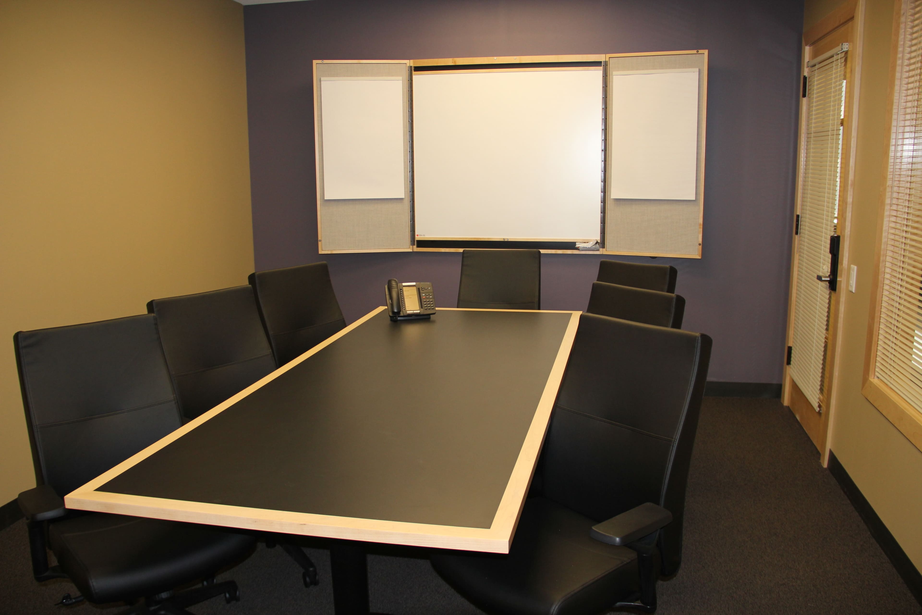 A conference room features a large black table surrounded by eight black chairs, with a whiteboard and door visible in the background.