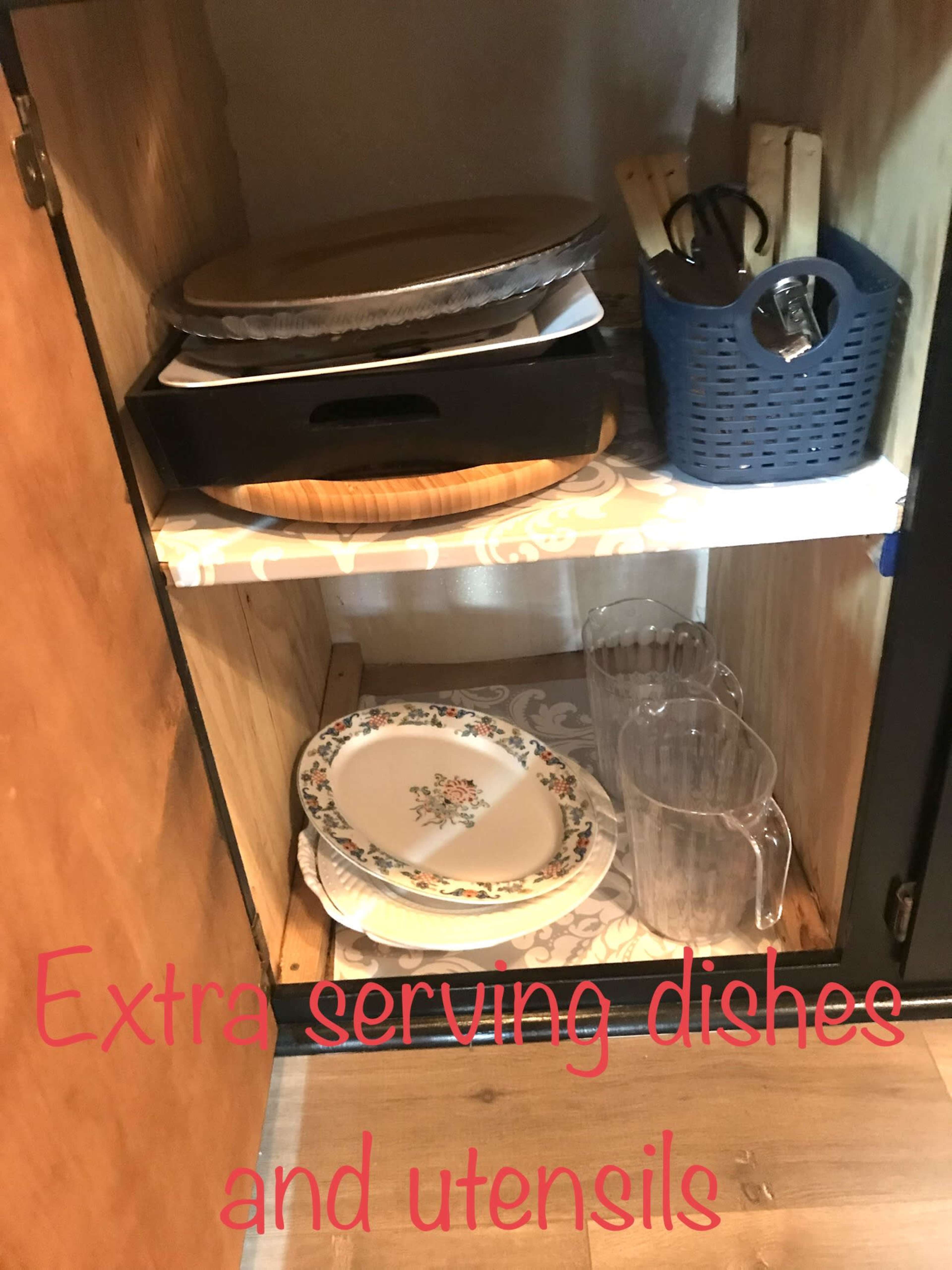 A wooden cabinet shelf holds a variety of serving dishes, utensils, and glass pitchers.