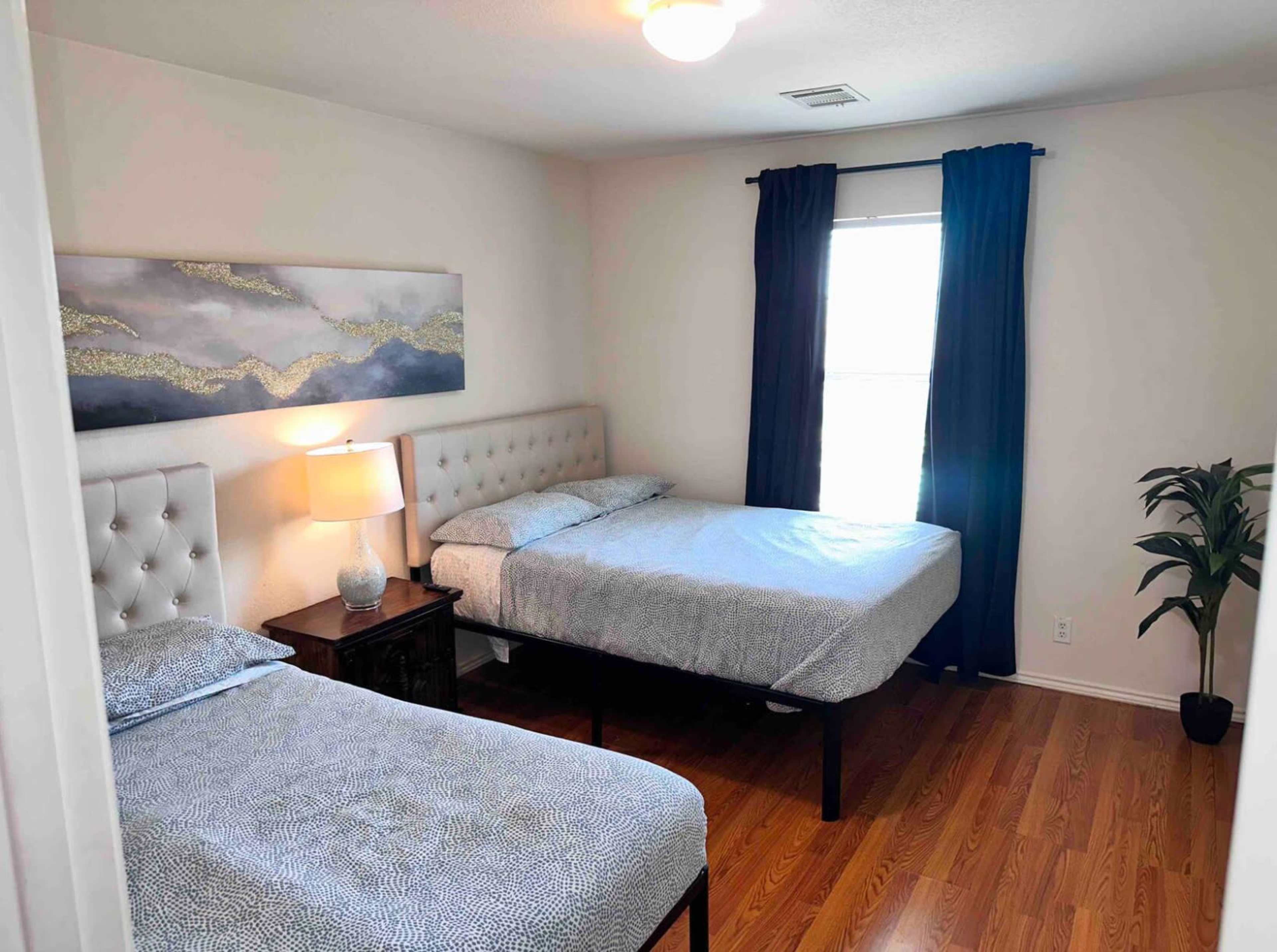 A small bedroom with two beds, a bedside table with a lamp, and a window covered by dark curtains.