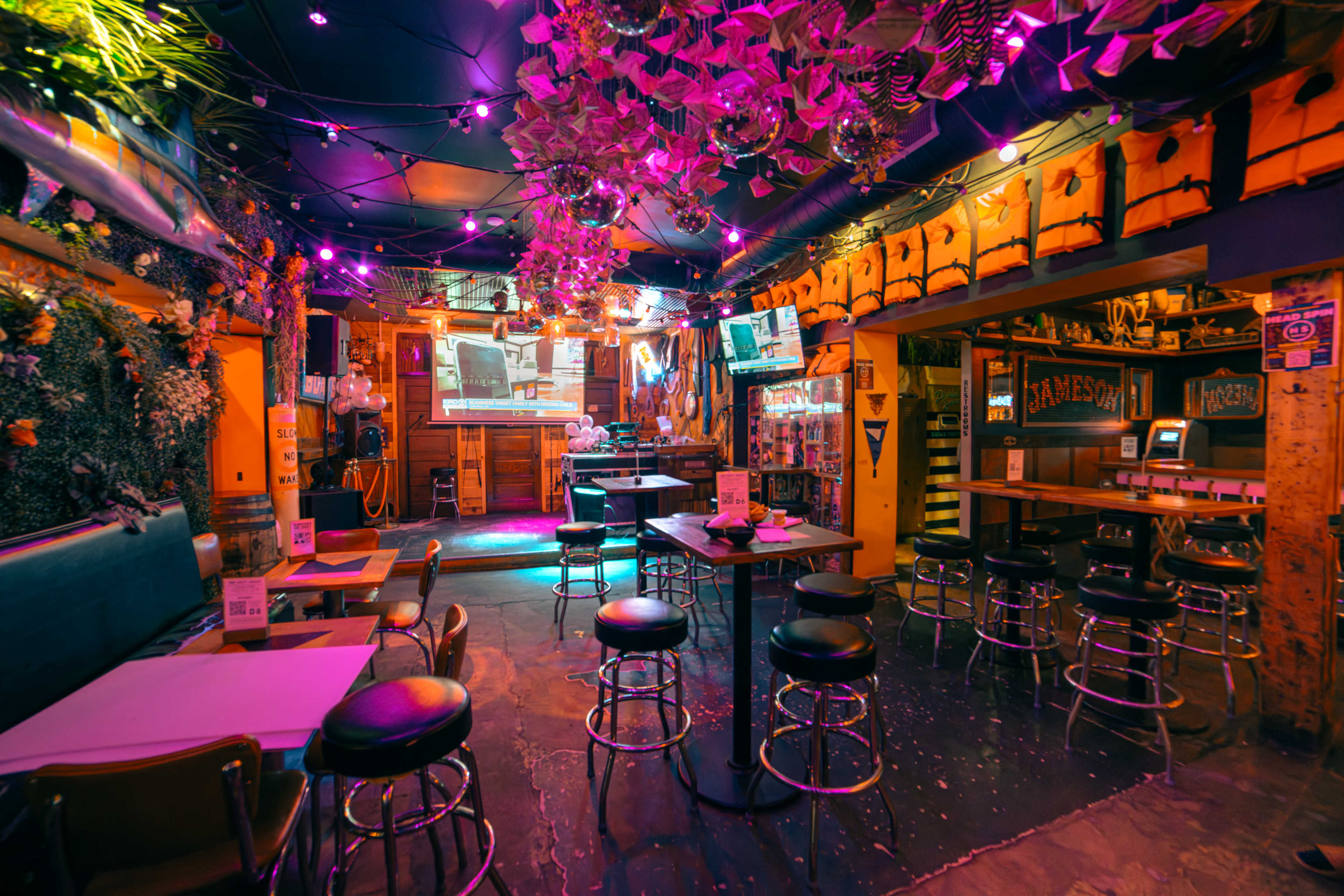 The image shows a vibrant bar interior with purple lighting, adorned with plants, displaying a projector screen at the back and a mix of high tables and stools.