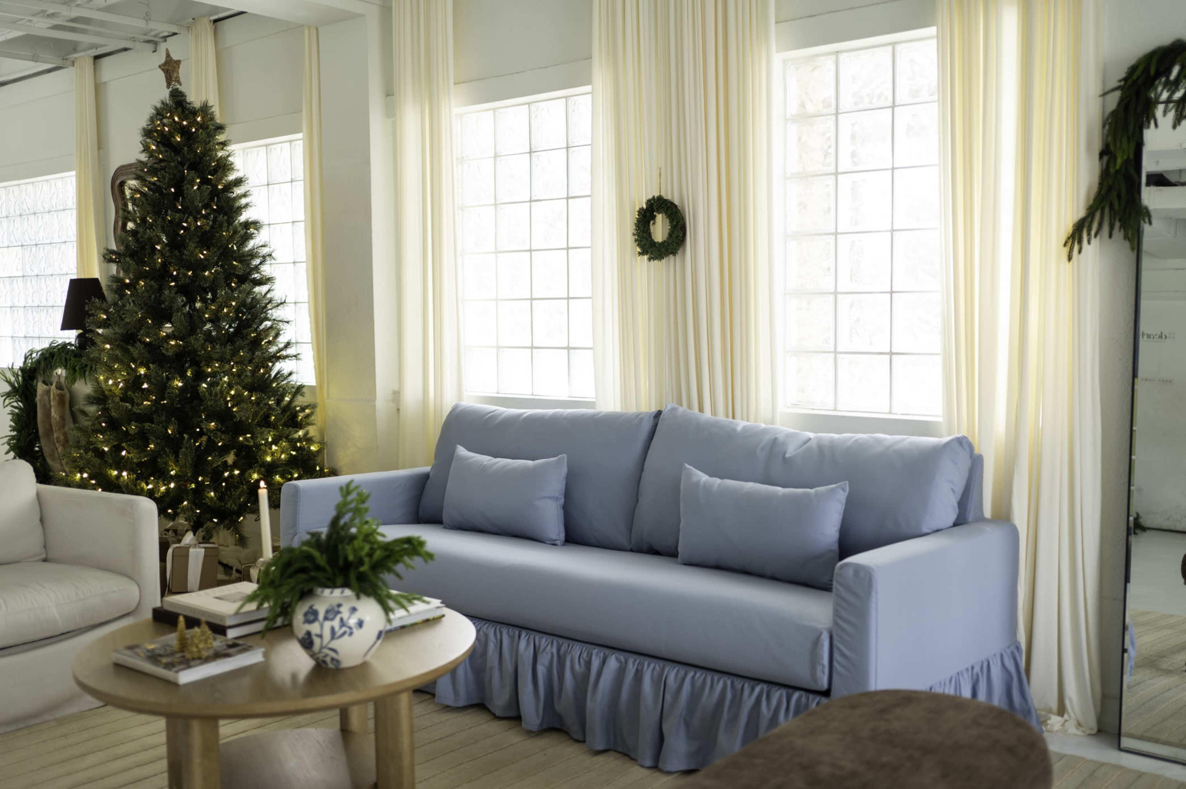 A light blue sofa with cushions is placed in a bright room decorated for the holidays, featuring a Christmas tree and large windows with sheer curtains.