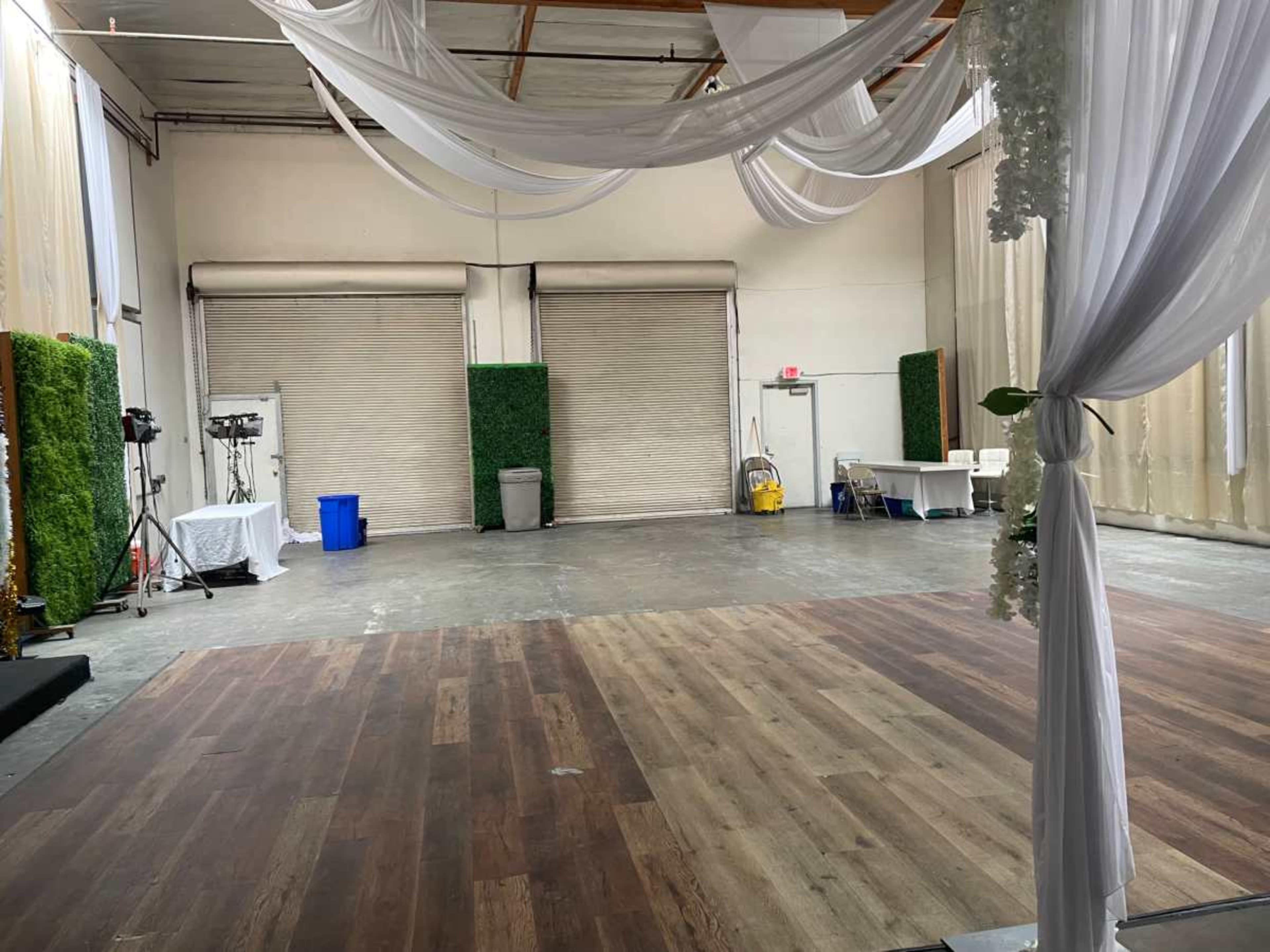 The image shows an empty event space with a wooden floor, draped fabric overhead, and large closed garage doors in the background.