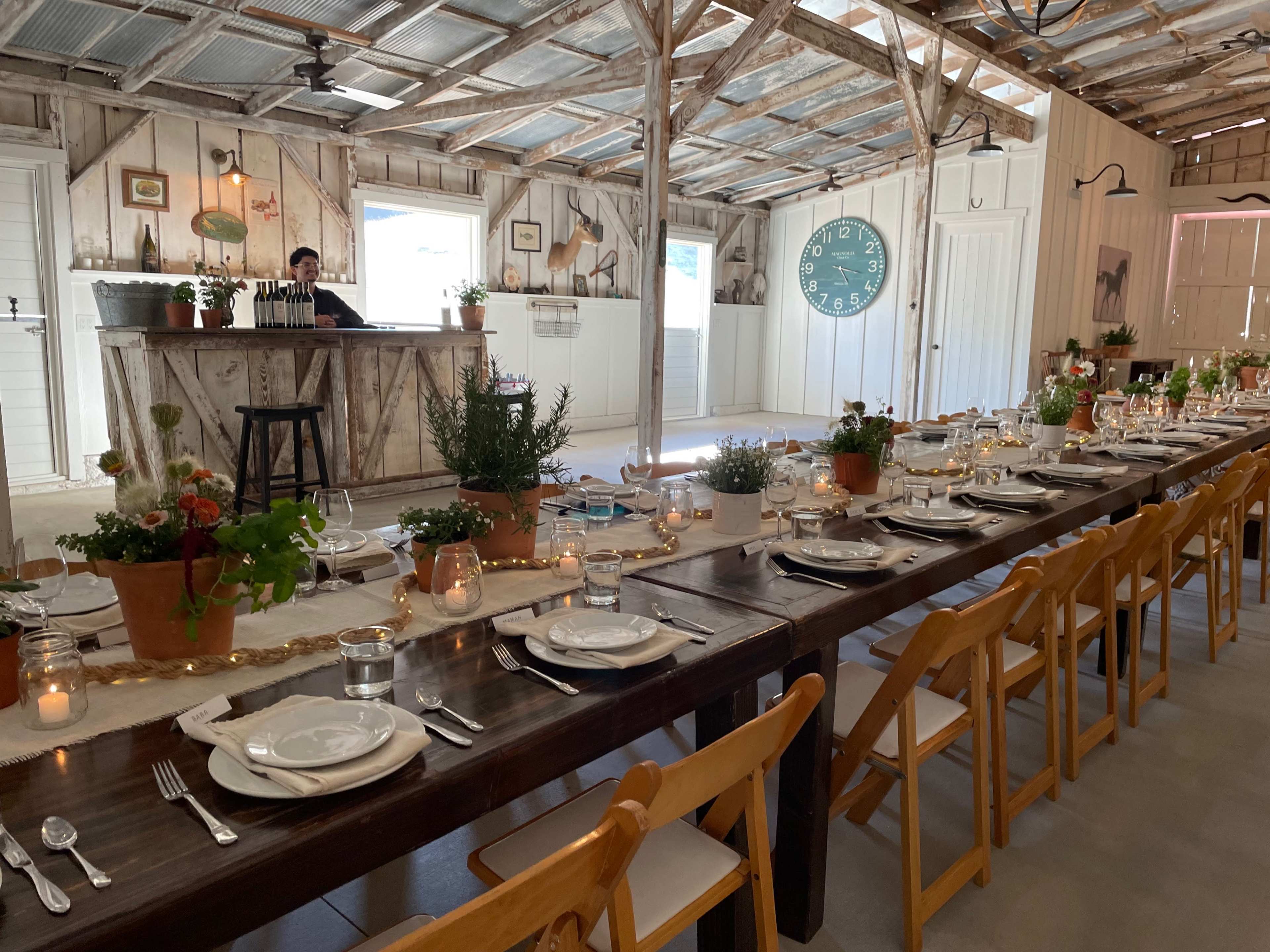 A long wooden table is set for a meal in a rustic indoor space featuring a bar, potted plants, and a large clock on the wall.