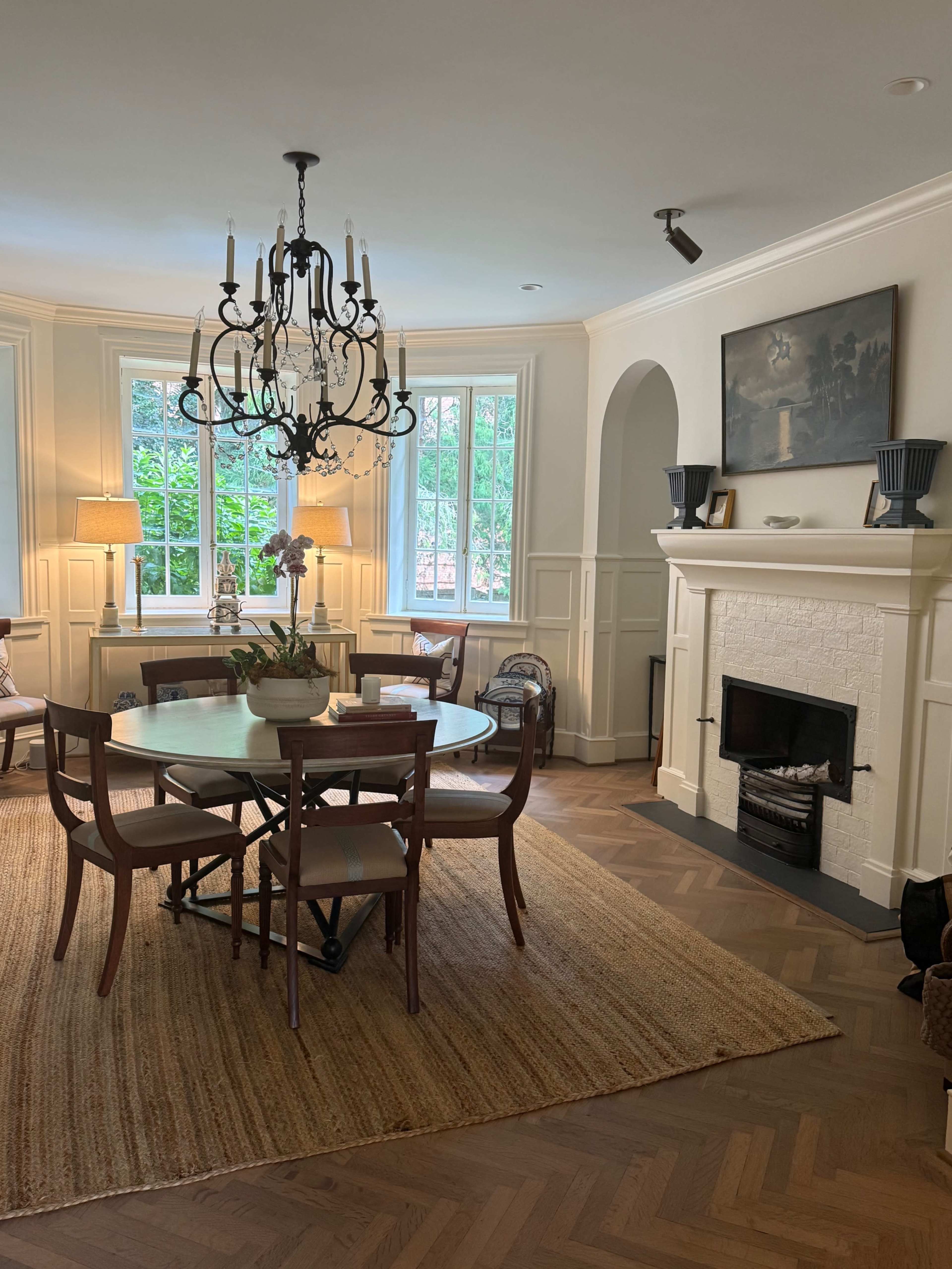 The image shows a well-lit dining area with a circular table surrounded by chairs, a decorative chandelier overhead, and a fireplace adorned with artwork and plants.