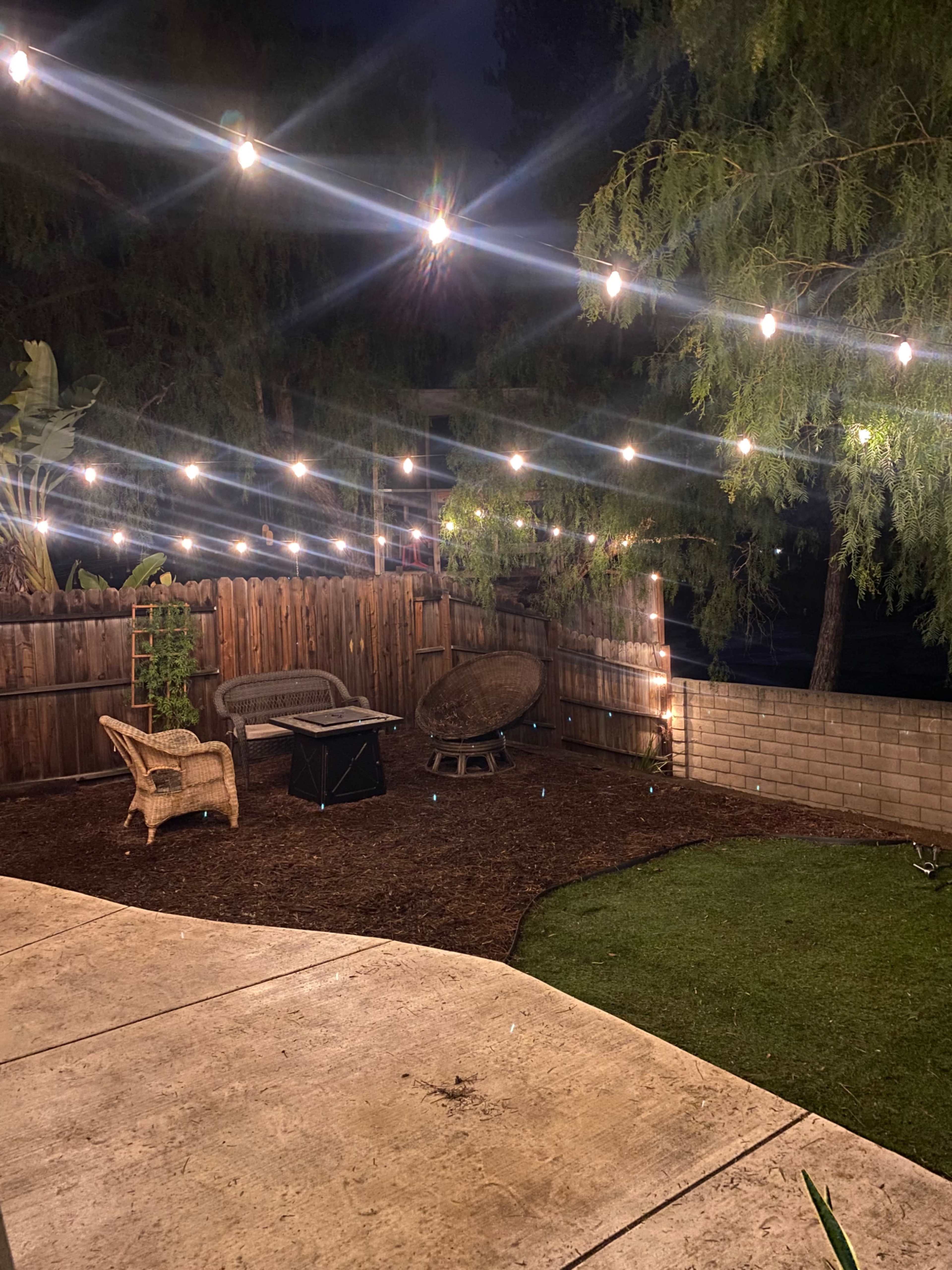 The image shows a backyard at night with string lights illuminating a seating area featuring wicker chairs and a small fire pit.
