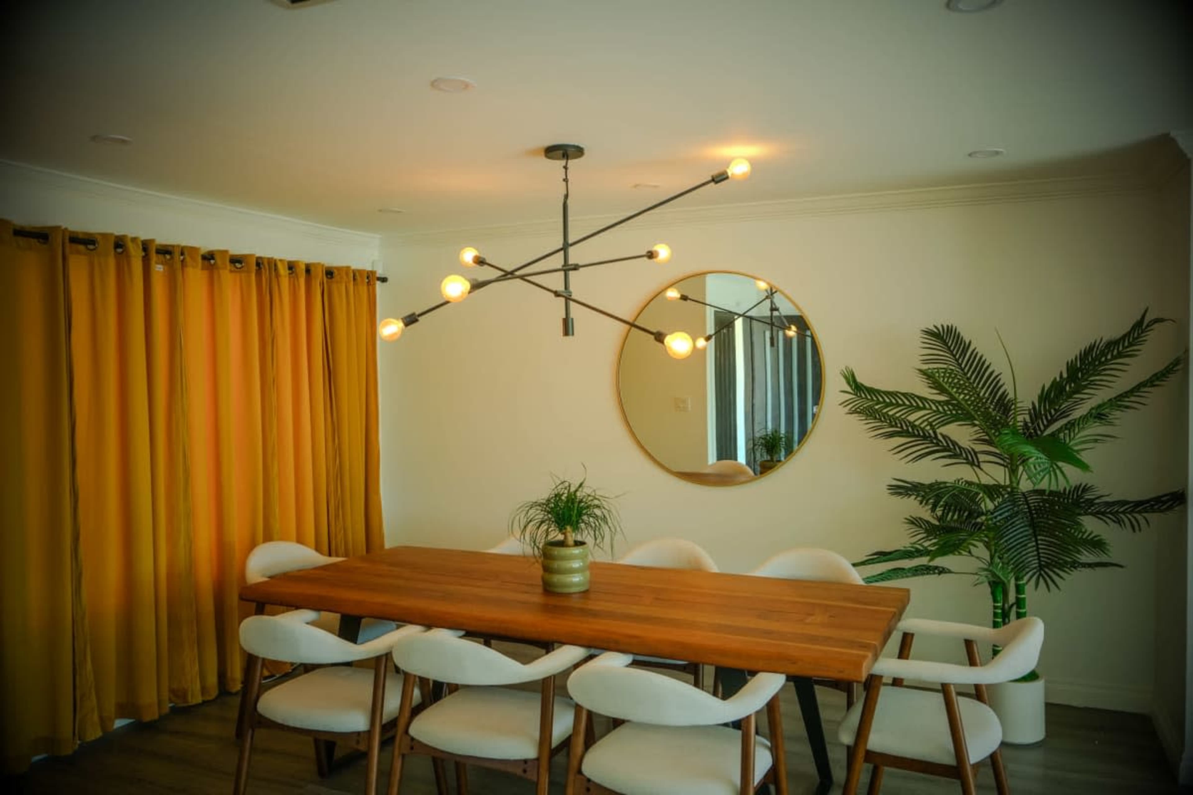 A modern dining room features a wooden table surrounded by white chairs, a large circular mirror on the wall, and yellow curtains draped by a window alongside a decorative plant.