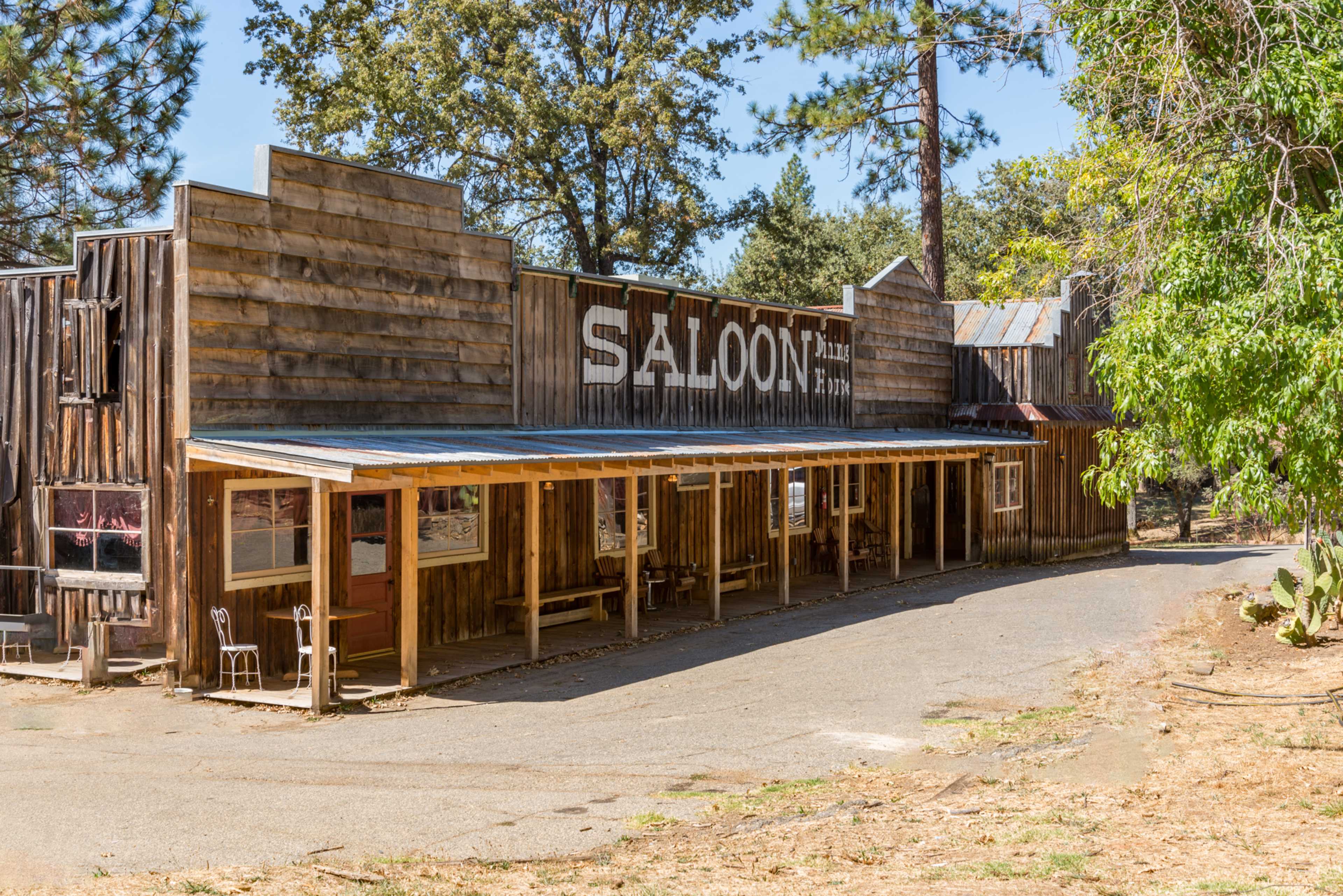A wooden saloon building with a covered porch is located along a winding path surrounded by trees.