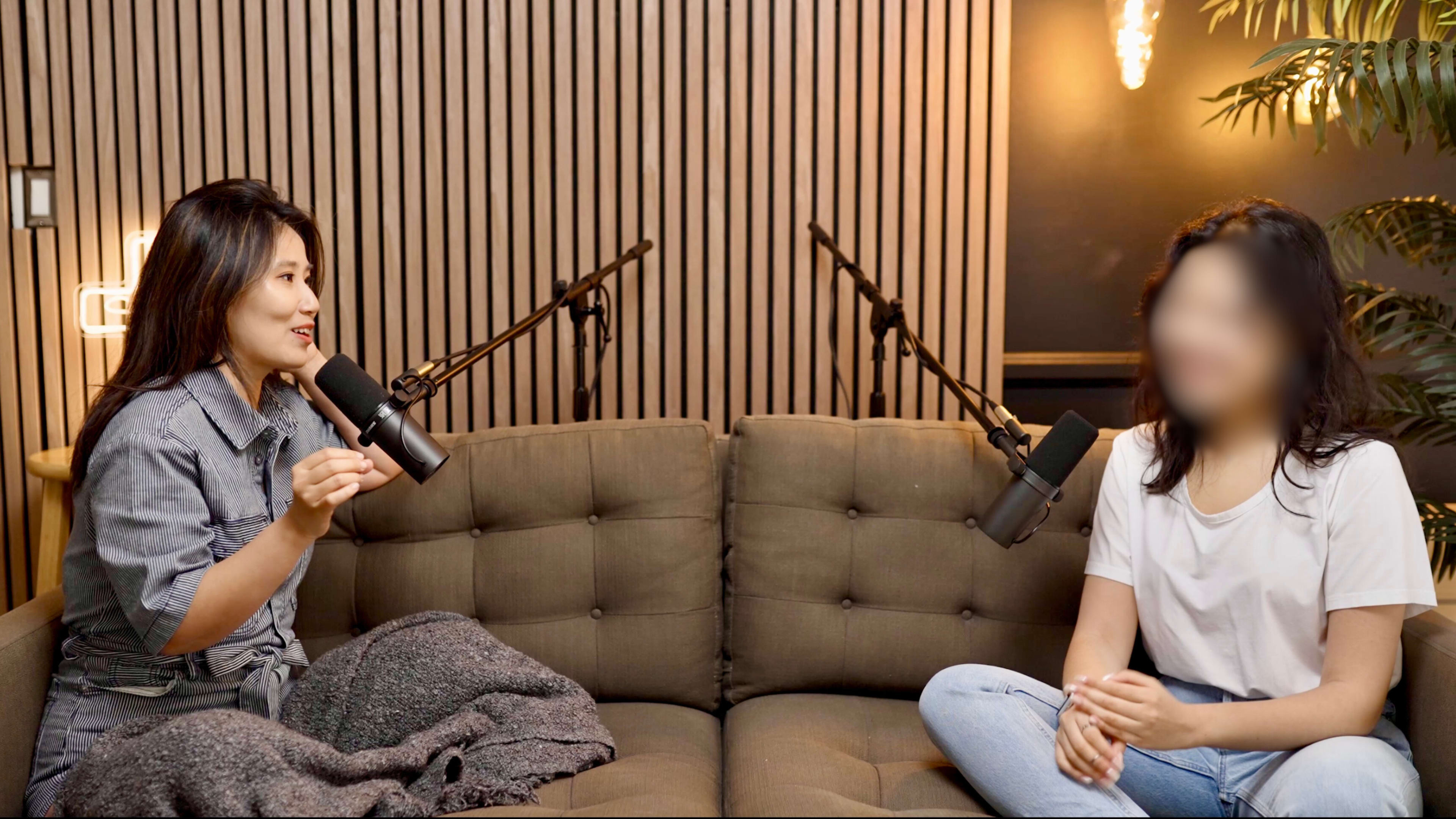 Two women are seated on a couch, each holding a microphone, in a studio setting with wooden paneling and microphone stands in the background.