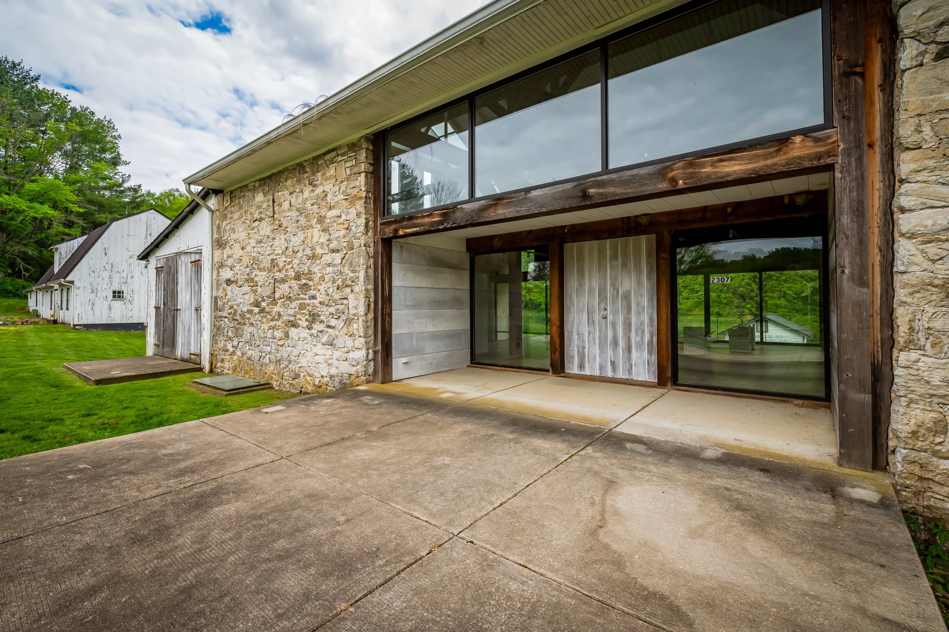 The image shows a modern building with large glass windows and a stone exterior, situated next to rustic barn-like structures on a grassy area.