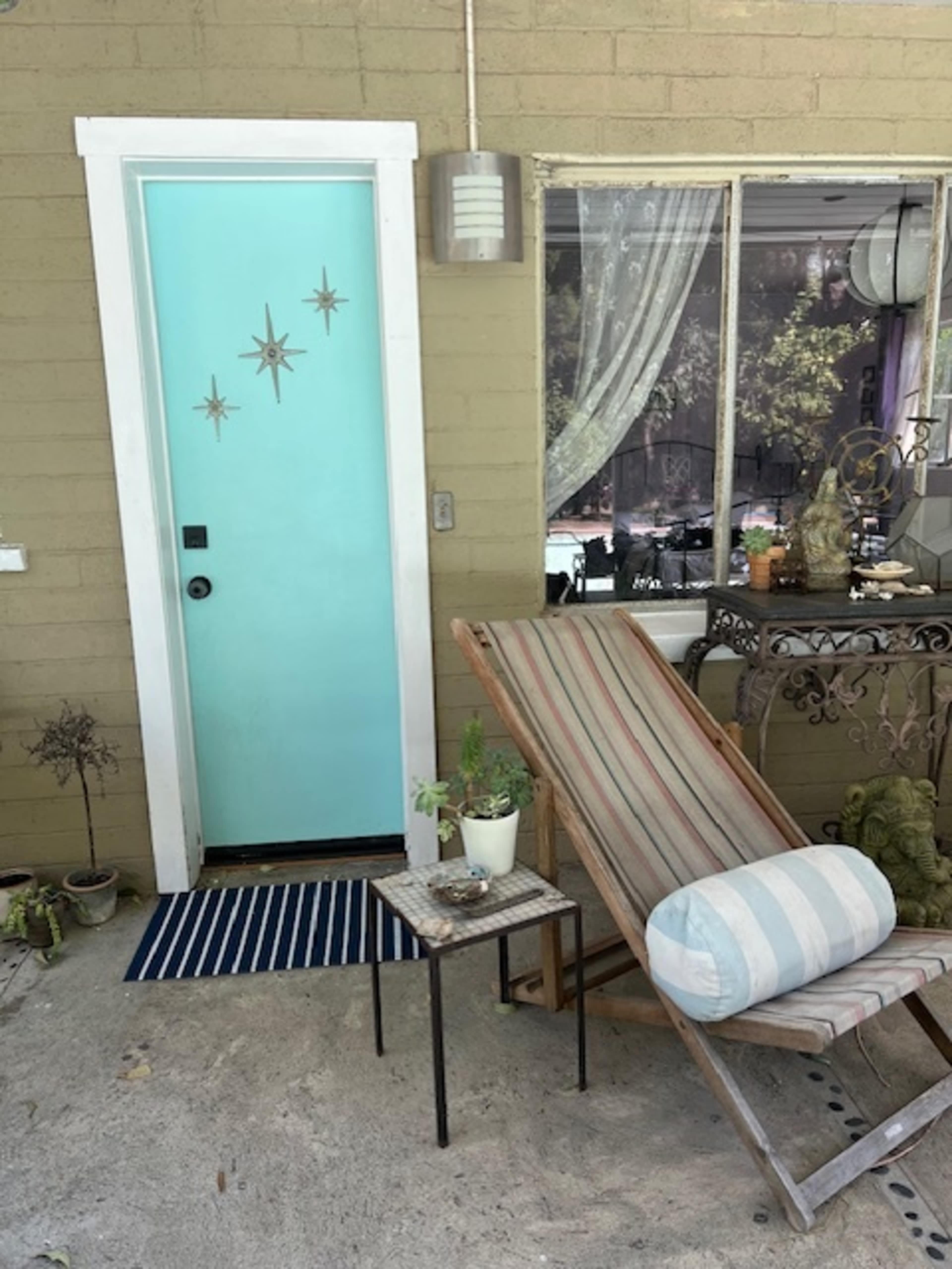 A turquoise door with star-shaped decorations stands next to a striped lounge chair and a small table with a potted plant.