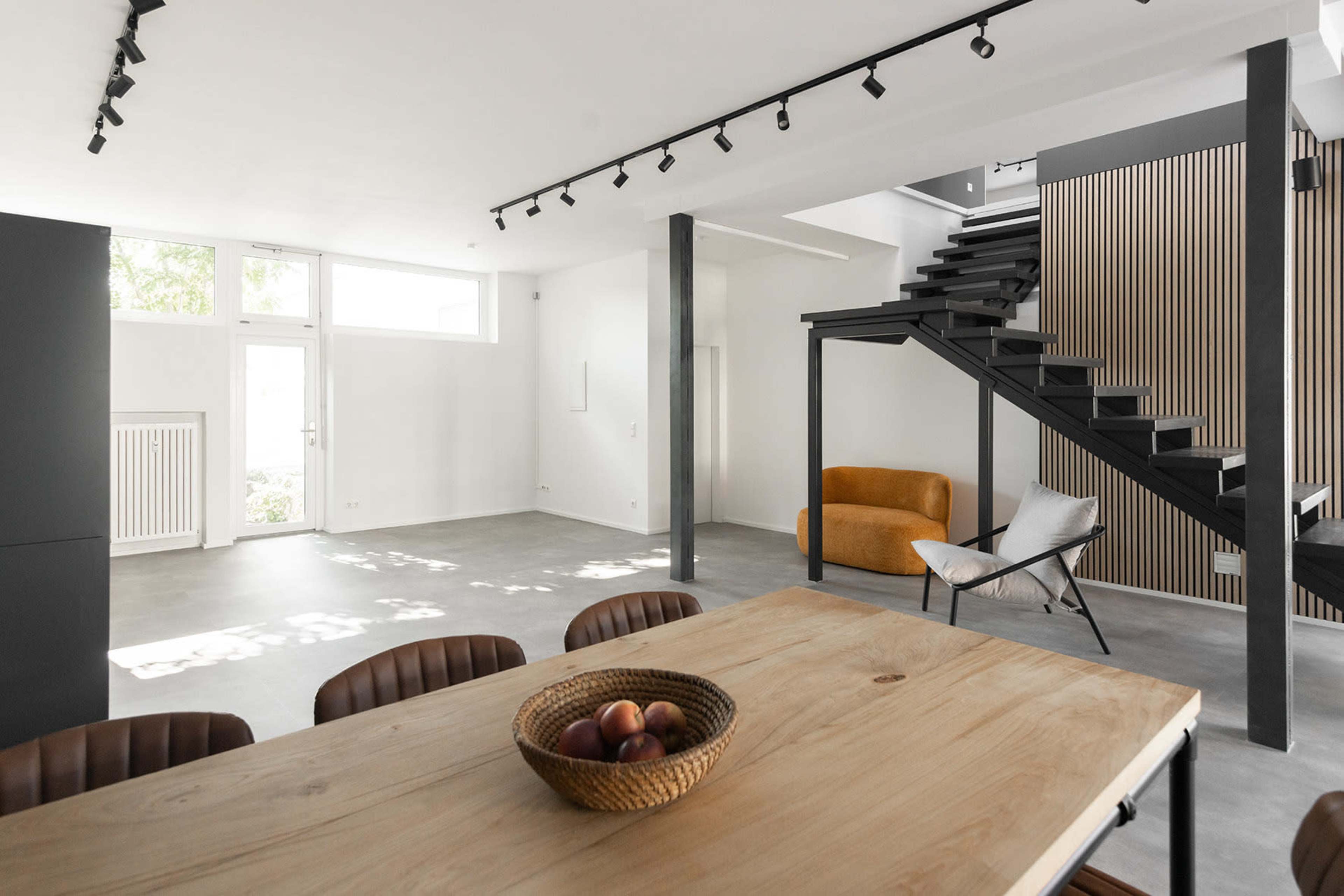 The image shows a modern, open-concept living space with a wooden dining table, a staircase, and minimalistic furniture.