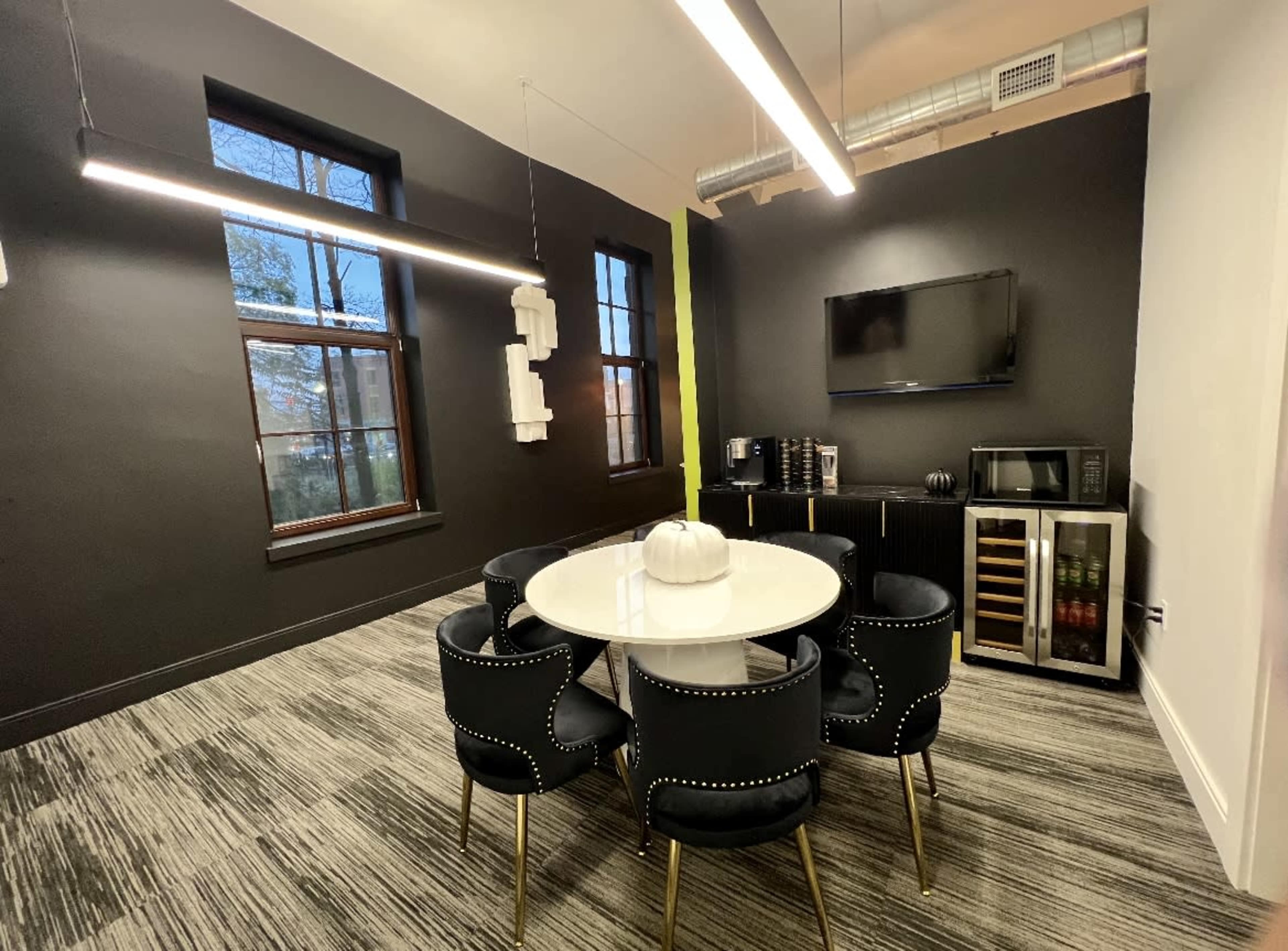 The image shows a modern meeting room featuring a round white table surrounded by black chairs, a wall-mounted TV, and a kitchenette area with a microwave and beverage cooler.