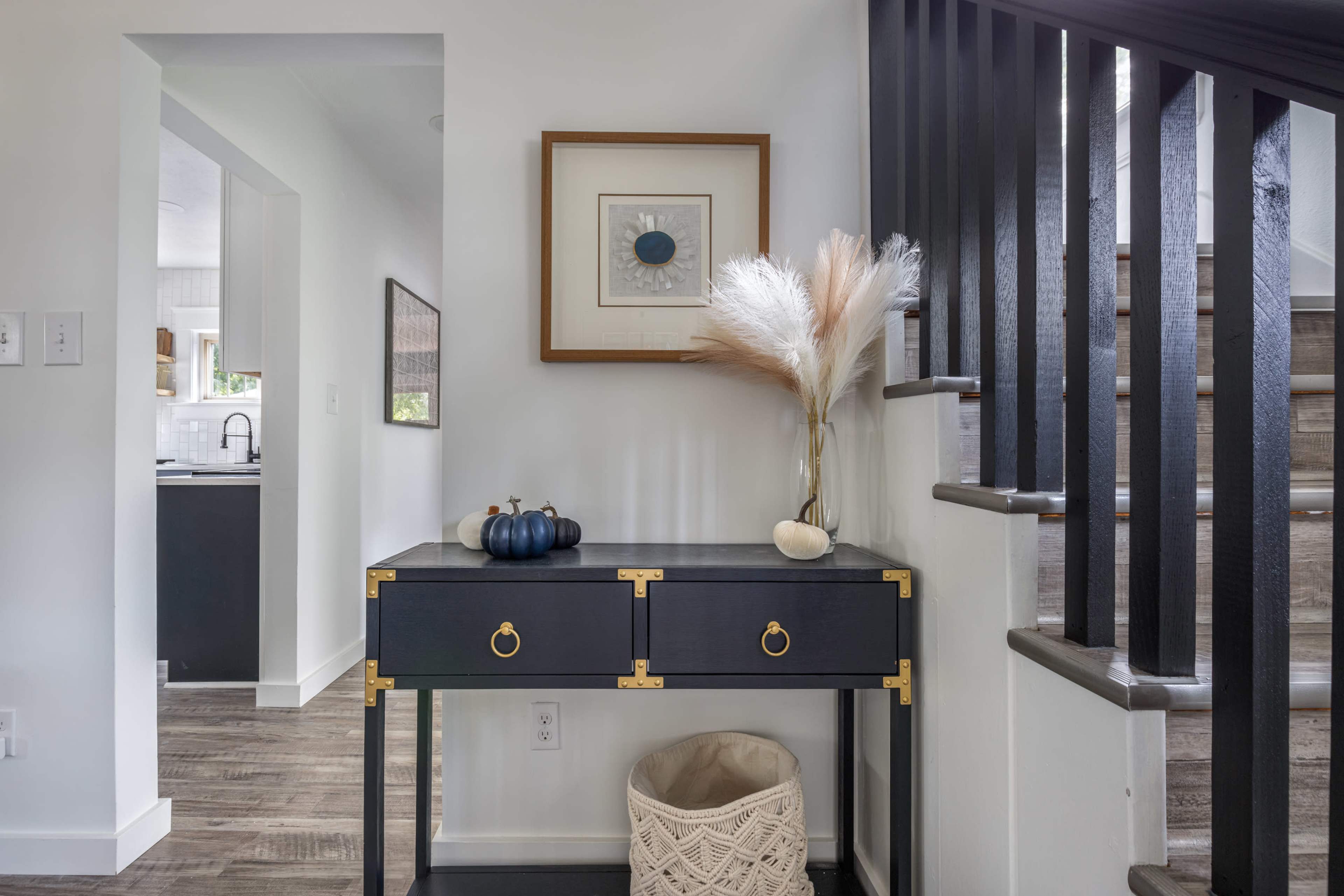 A black console table with two drawers, decorated with a framed artwork and a vase of dried flowers, is positioned beside a staircase in a bright hallway.
