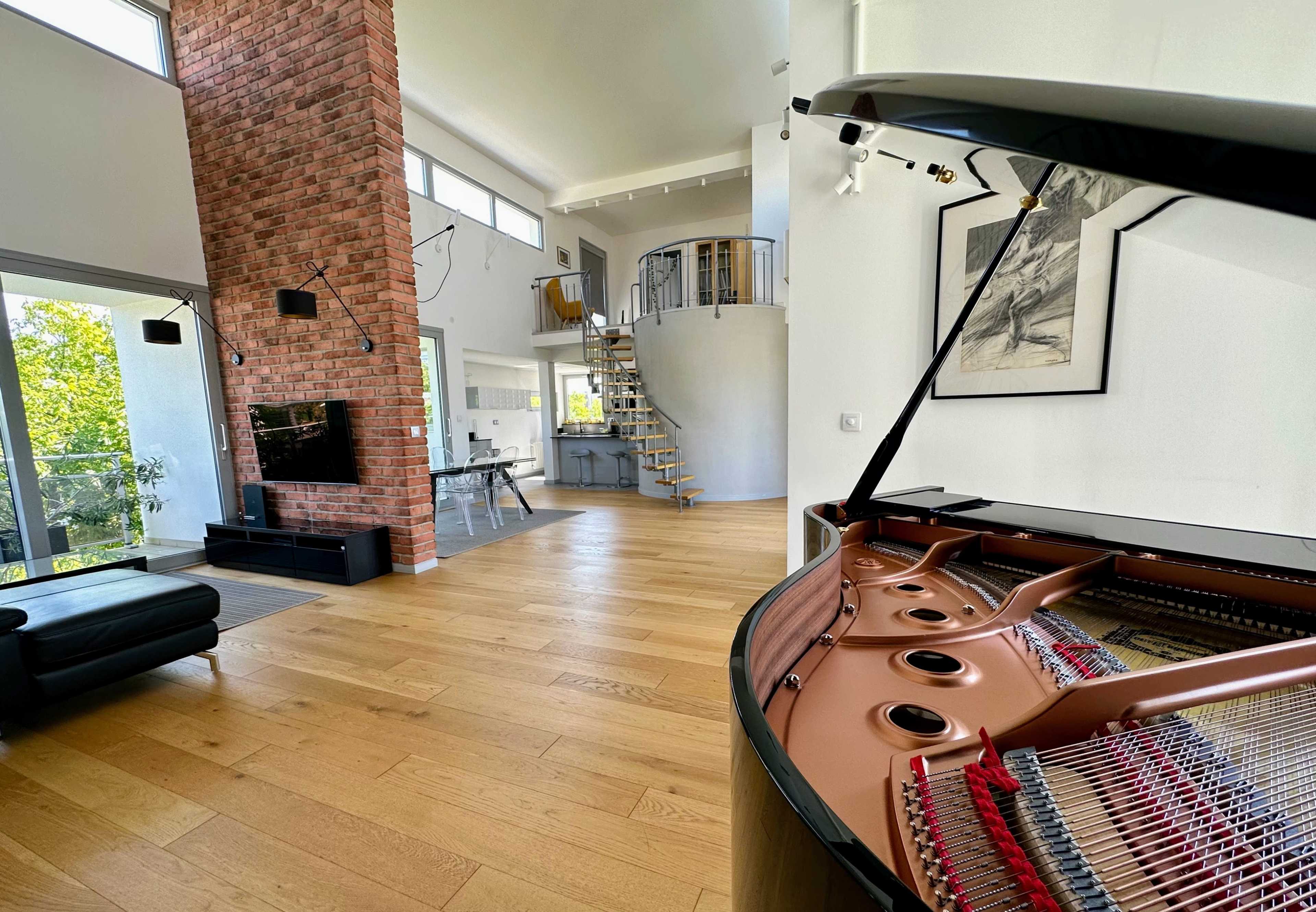 An elegant living space features a grand piano in the foreground, with a high ceiling, a brick wall, and a spiral staircase in the background.