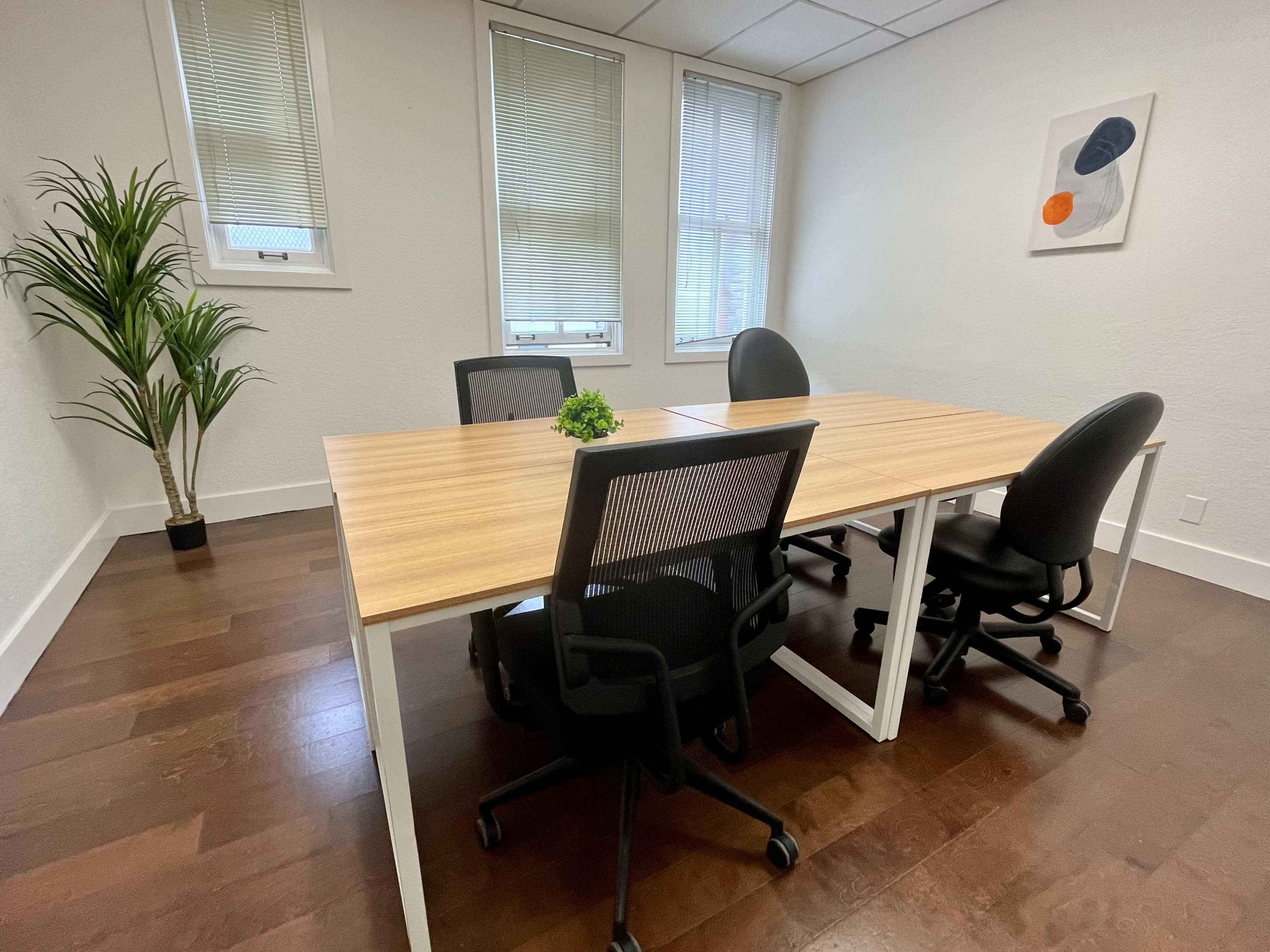 A modern office meeting room features a wooden table surrounded by four black chairs, with a potted plant in one corner and a piece of artwork on the wall.