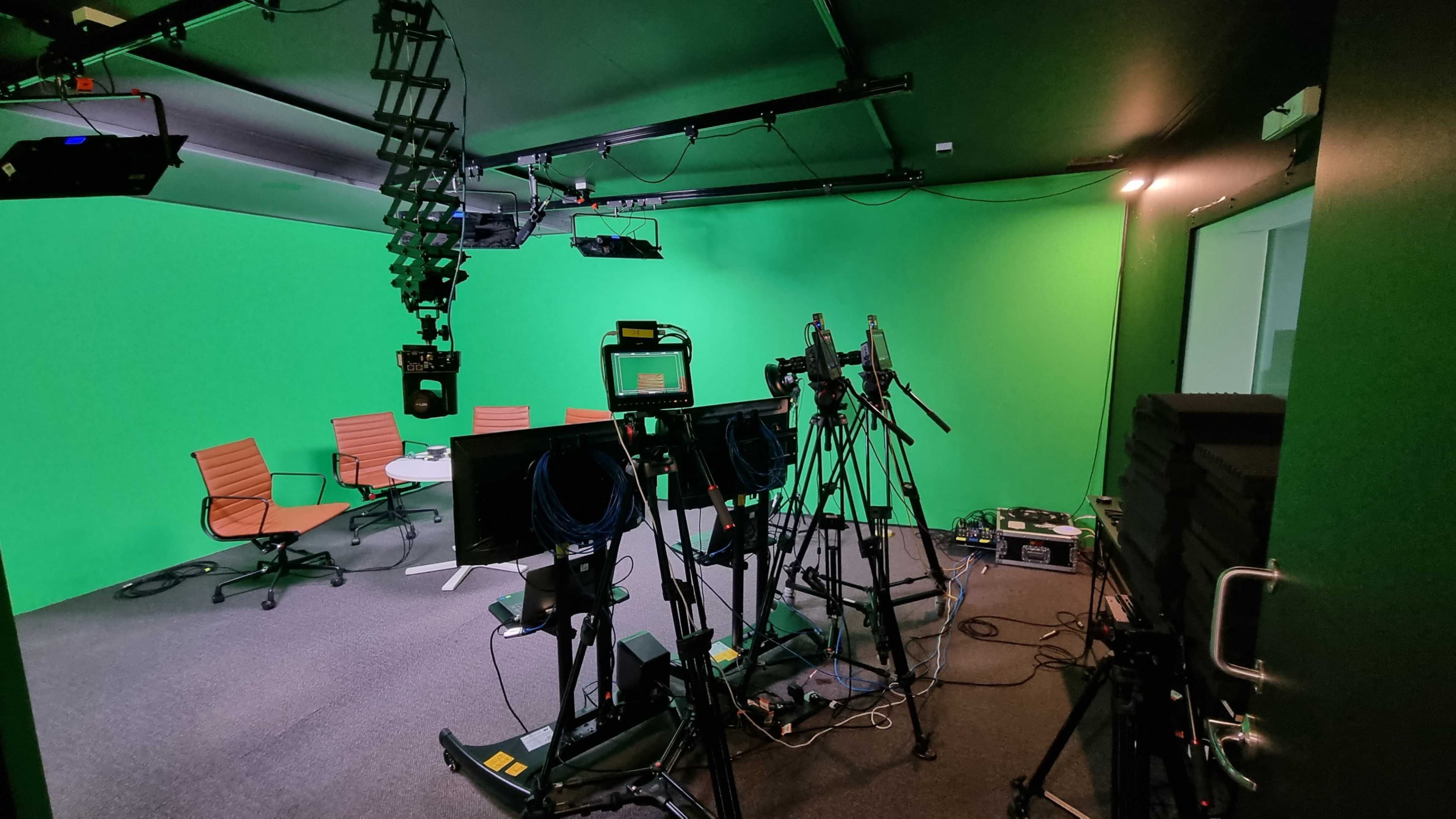 The image shows a production studio featuring a green screen, empty chairs and table, and various cameras and equipment set up for filming.