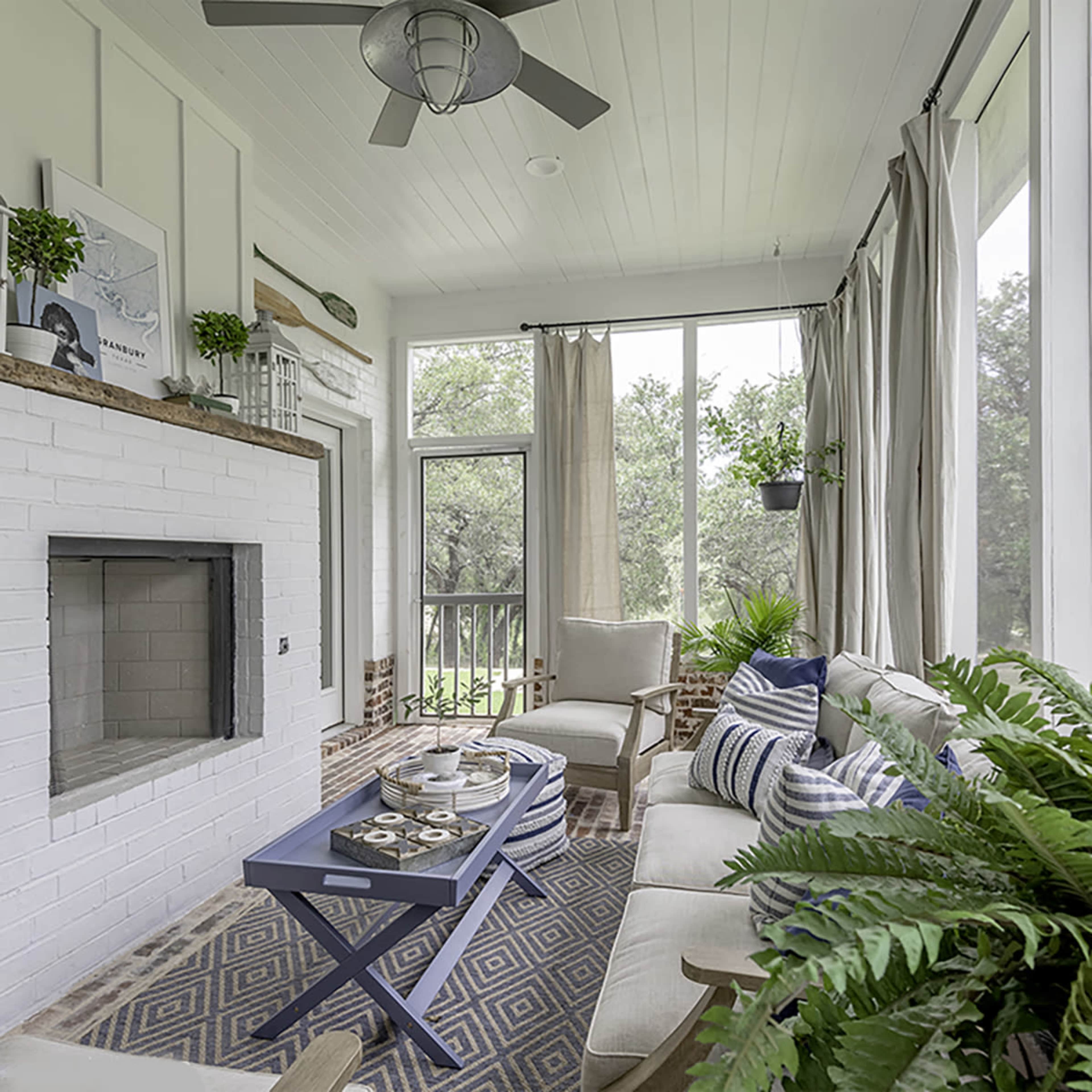 A sunlit sunroom with a fireplace, comfortable seating, a blue coffee table, and potted plants lining the windows.