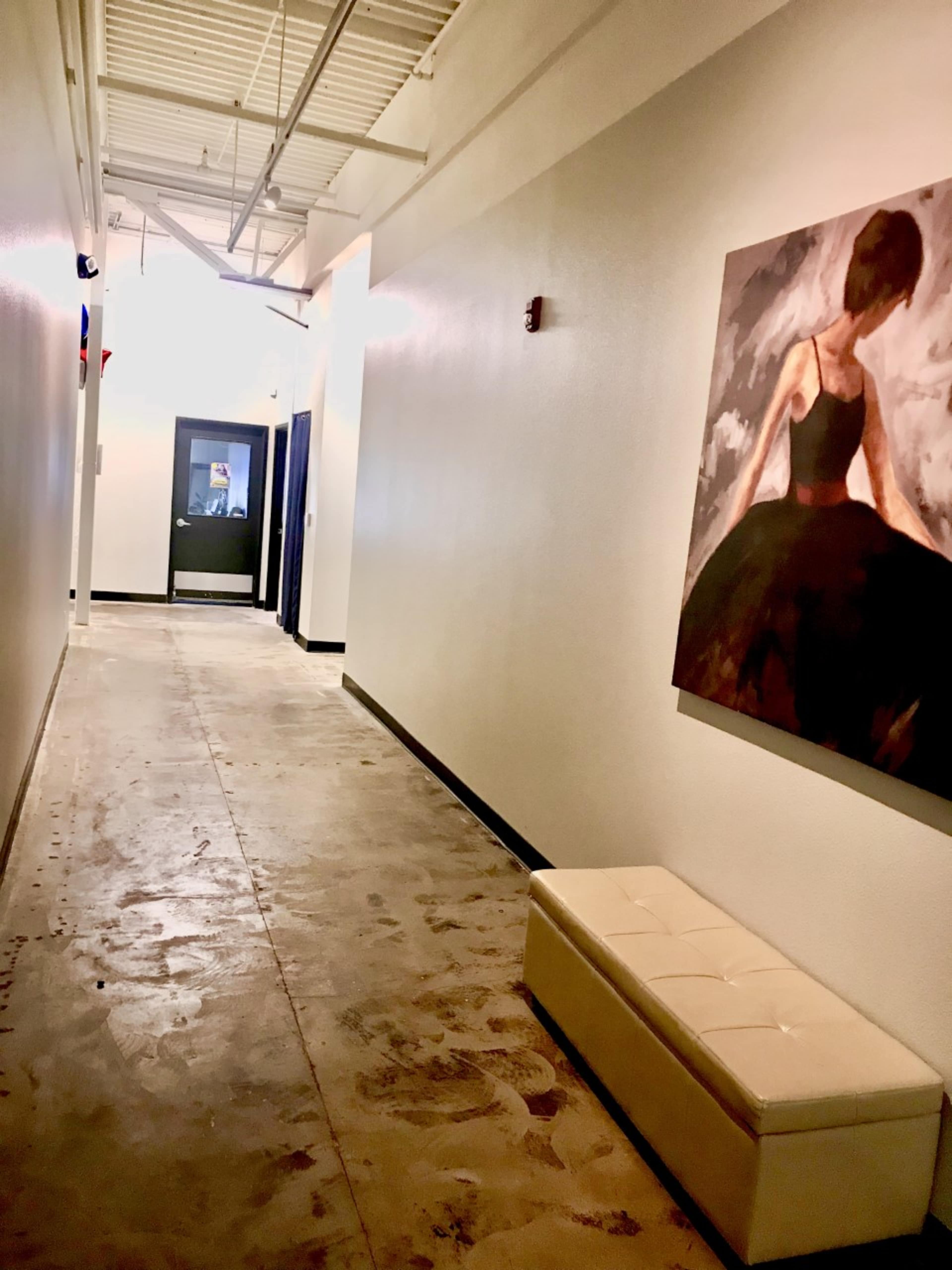 A narrow hallway with a textured concrete floor, a white wall featuring a painting of a woman in a black dress, and a small white bench on the side.