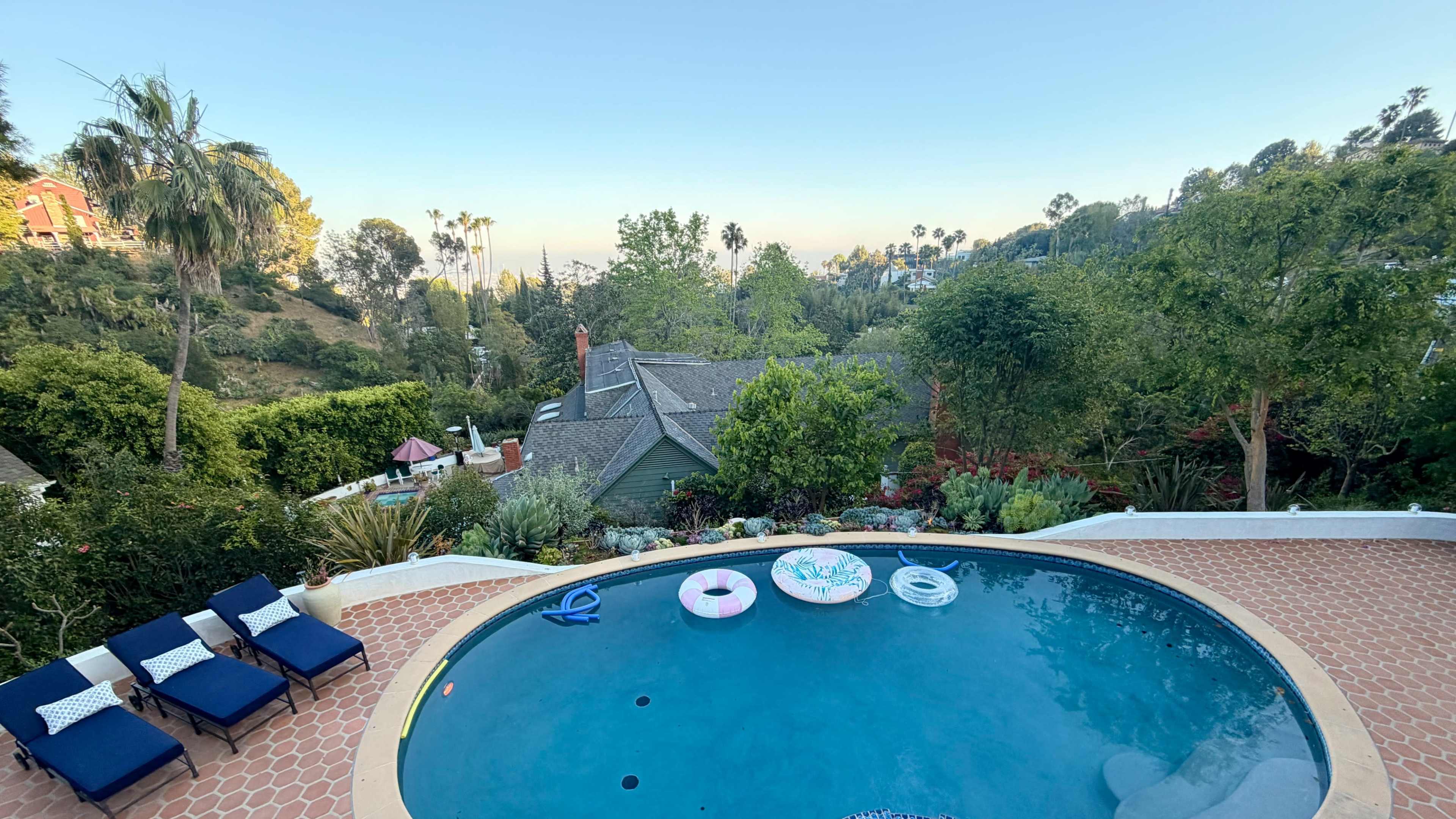 The image shows a swimming pool with lounge chairs and floaties, surrounded by lush vegetation and a hillside view.
